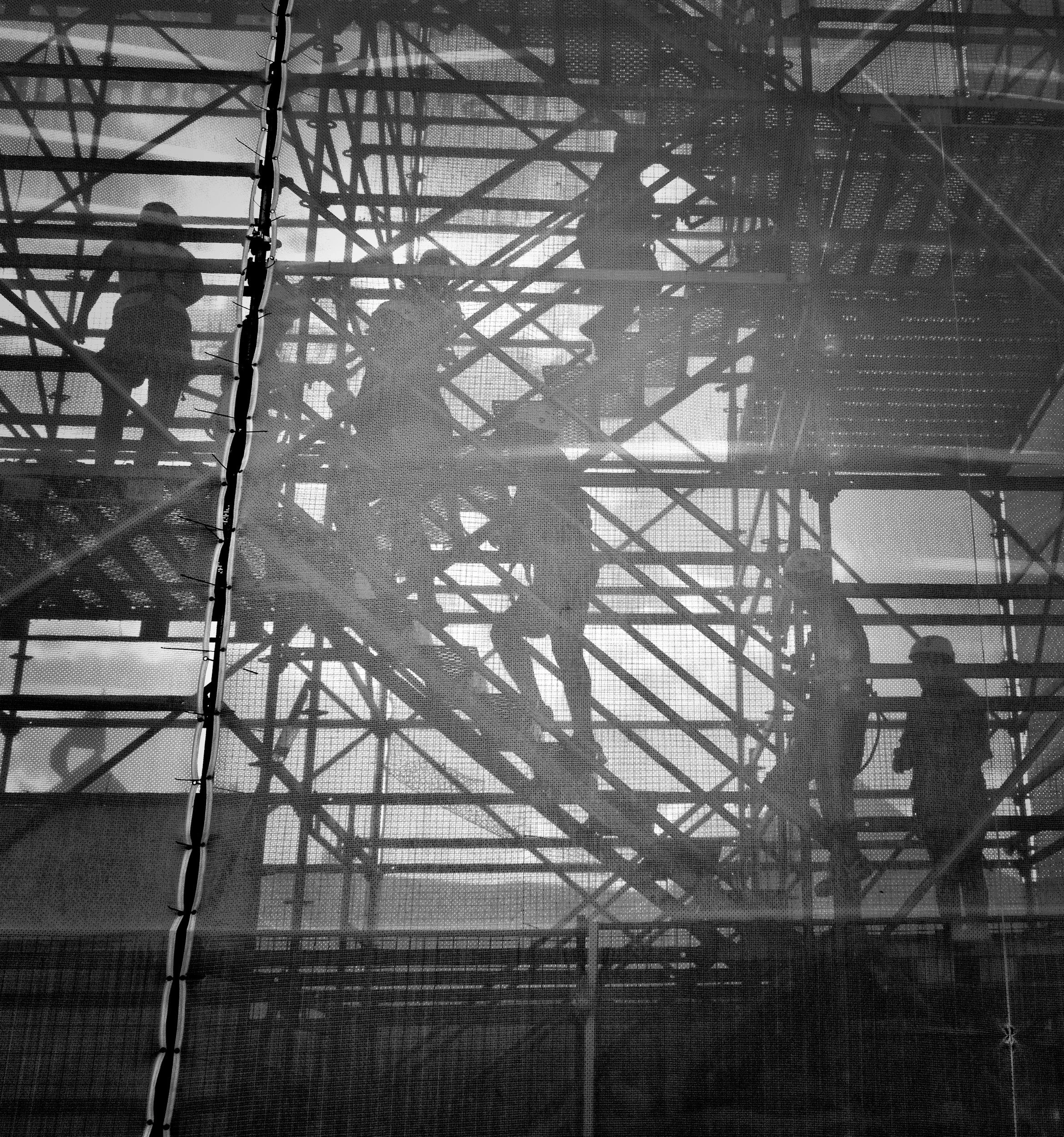 Black and white photo of construction workers and their shadows as they climb metal, temporary stairs to access an elevated worksite.