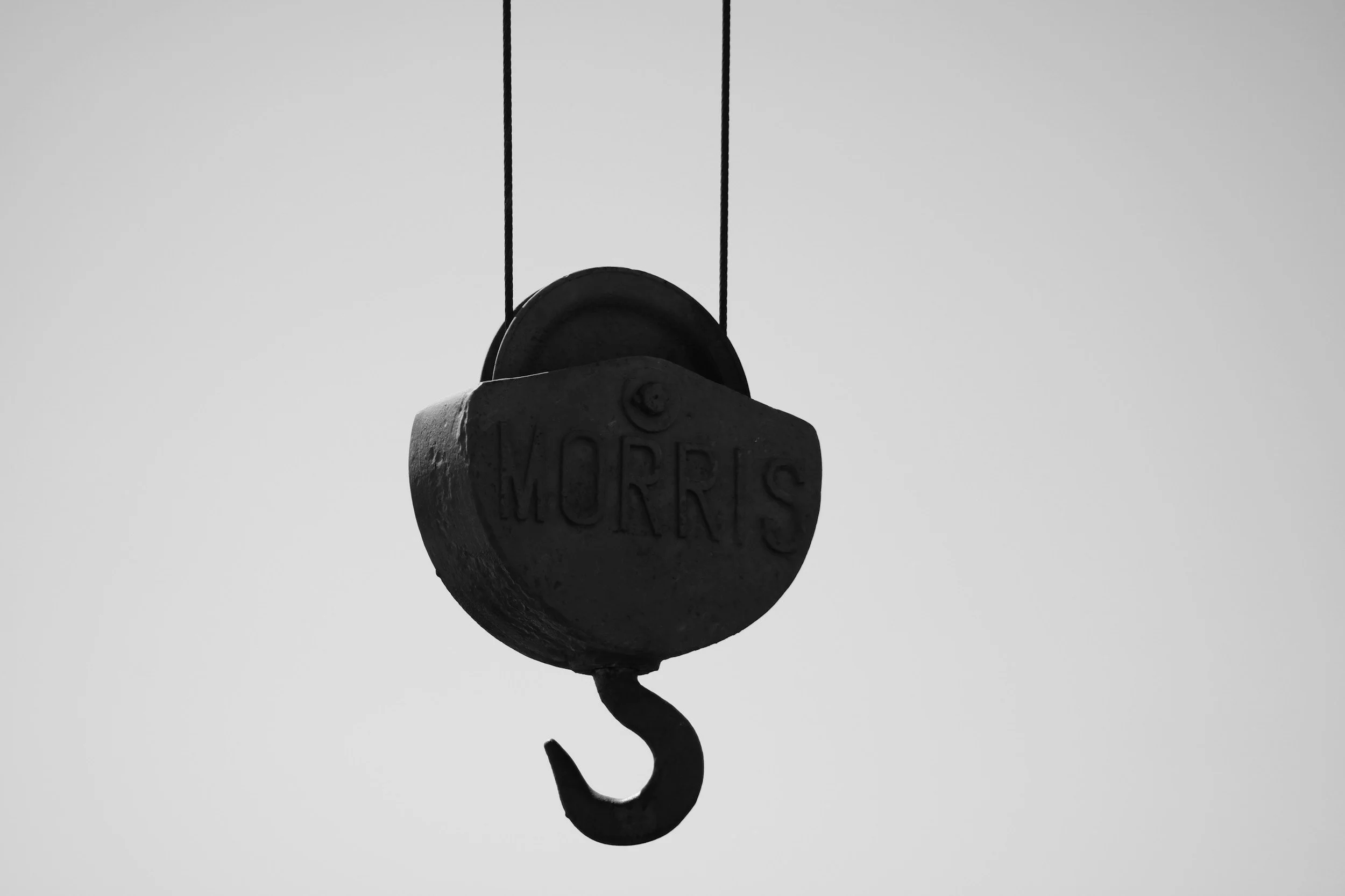 Close-up of an old mechanical hoist with hook with the word 'MORRIS' embossed on it, hanging against a plain sky background.