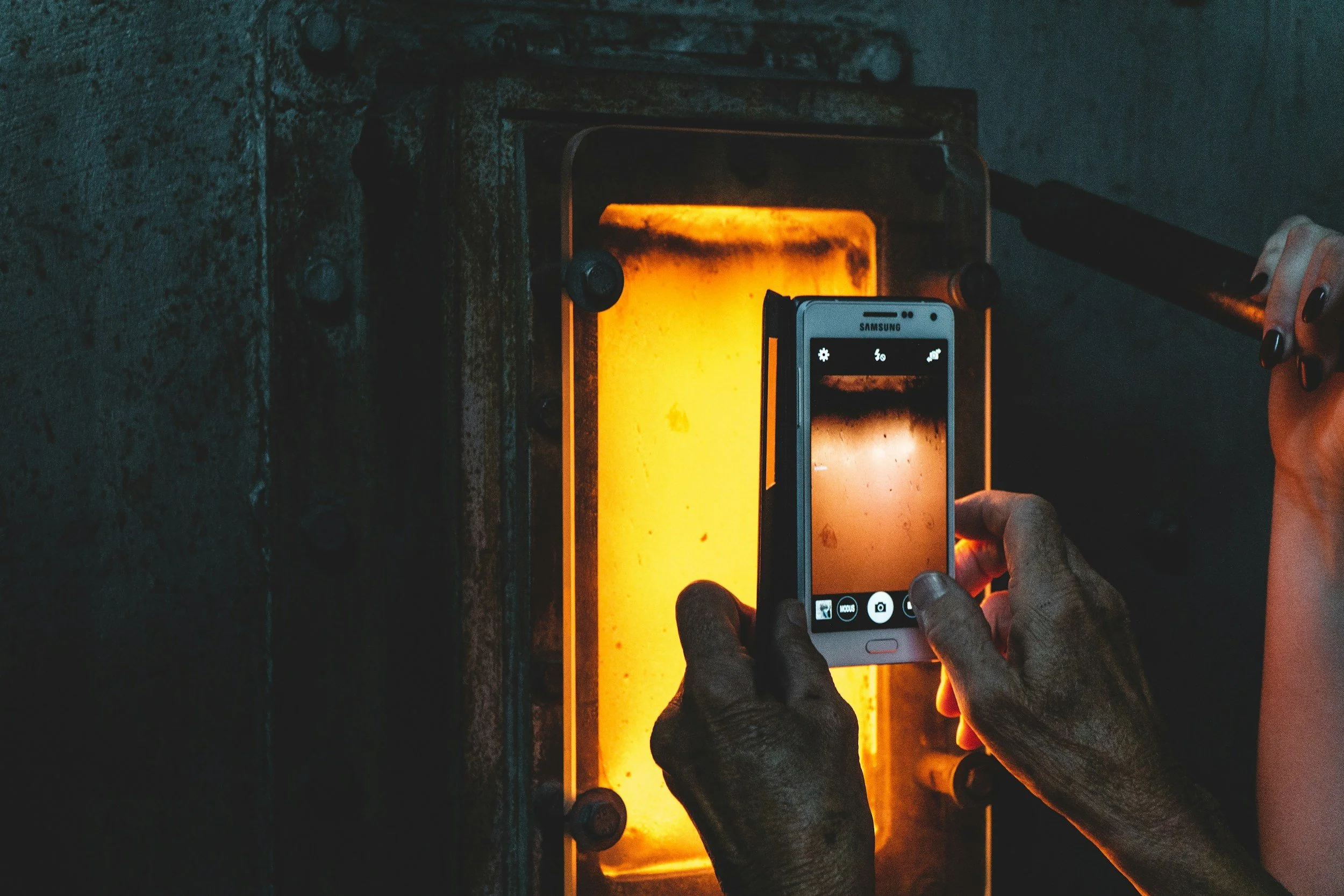 A person is taking a photo with a Samsung smartphone of a glowing orange rectangular furnace or kiln in a foundry, with a dark background and a hand gripping a tool to the right.