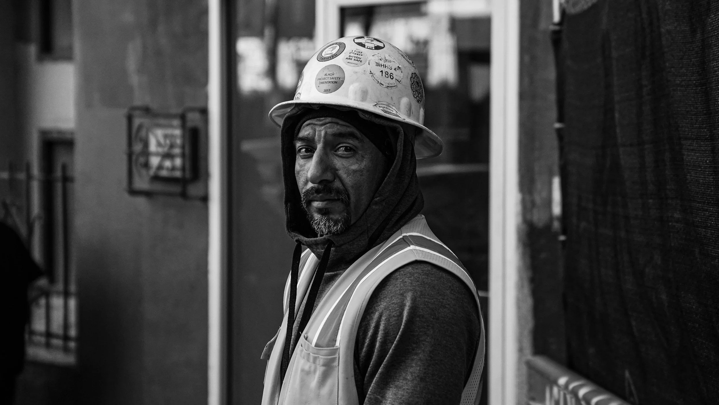 A construction worker with a beard, wearing a safety helmet and reflective vest, standing indoors with a serious expression on his face.