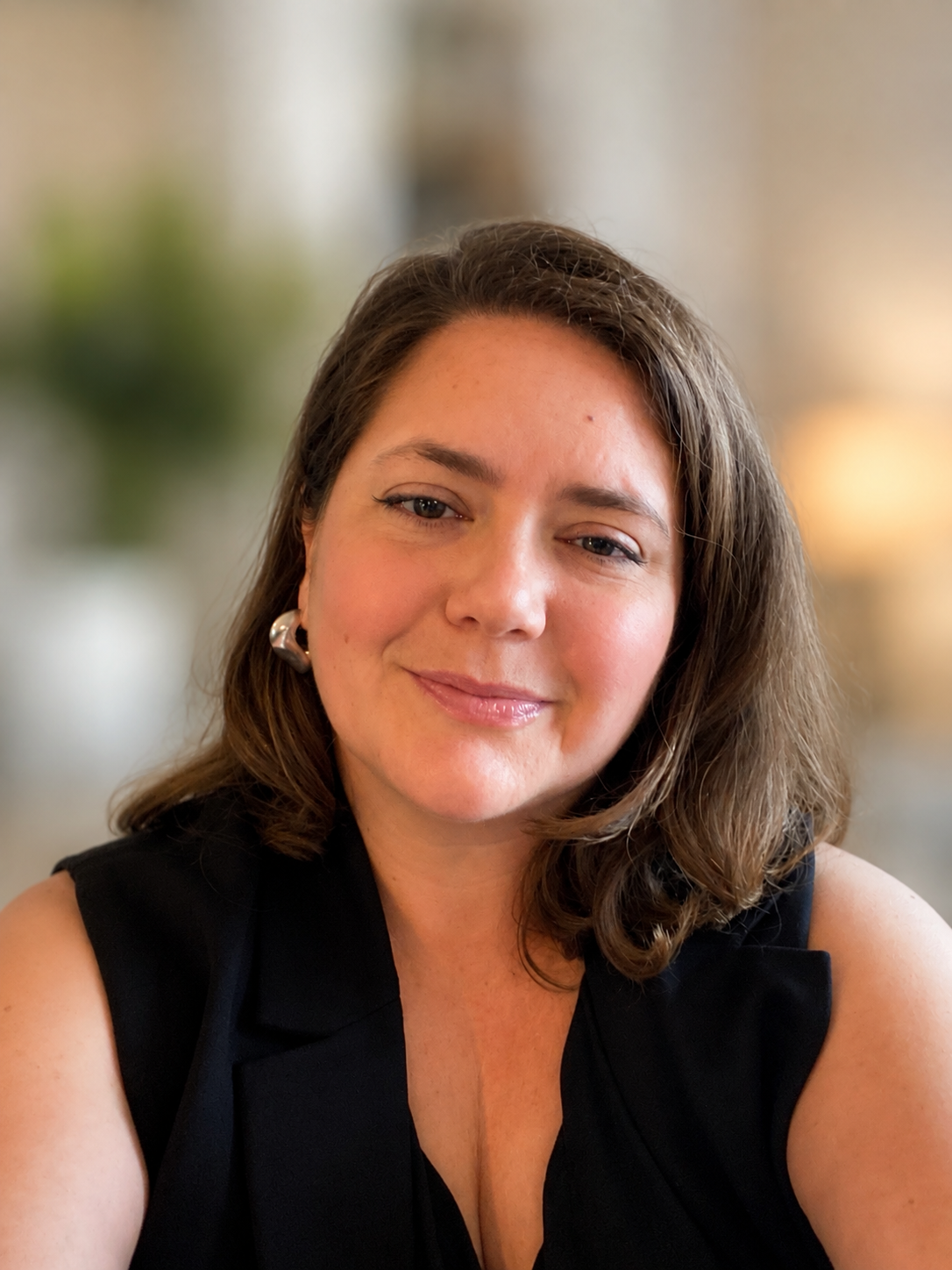 Hillary Waterhouse with shoulder-length brown hair, wearing a black sleeveless top and silver earrings, smiling at the camera with a blurred indoor background.