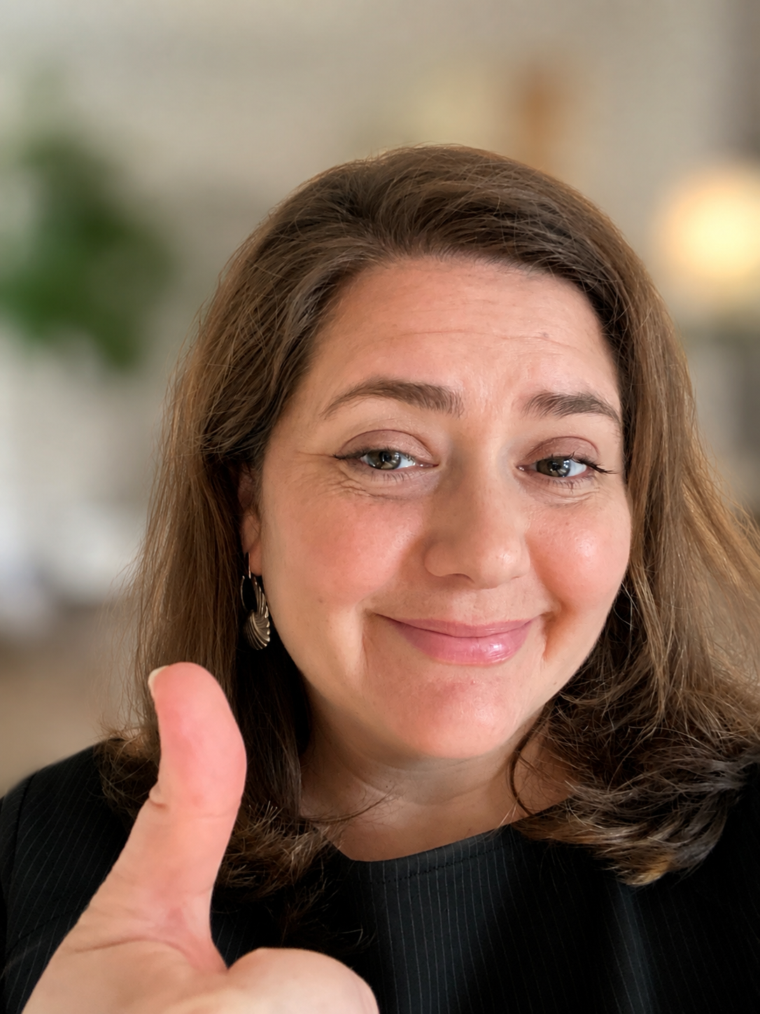 Hillary Waterhouse smiling and giving a thumbs-up gesture, with brown hair, earrings, and a black top, in a softly lit indoor setting.