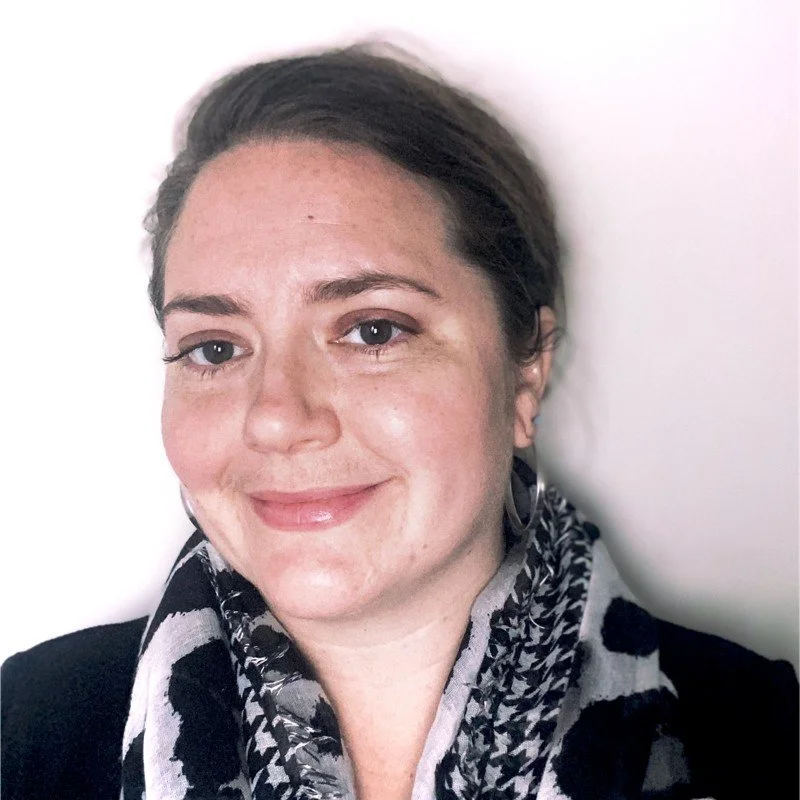A woman with dark blonde hair smiling, wearing a black and white patterned scarf and a black top, standing against a plain white wall.