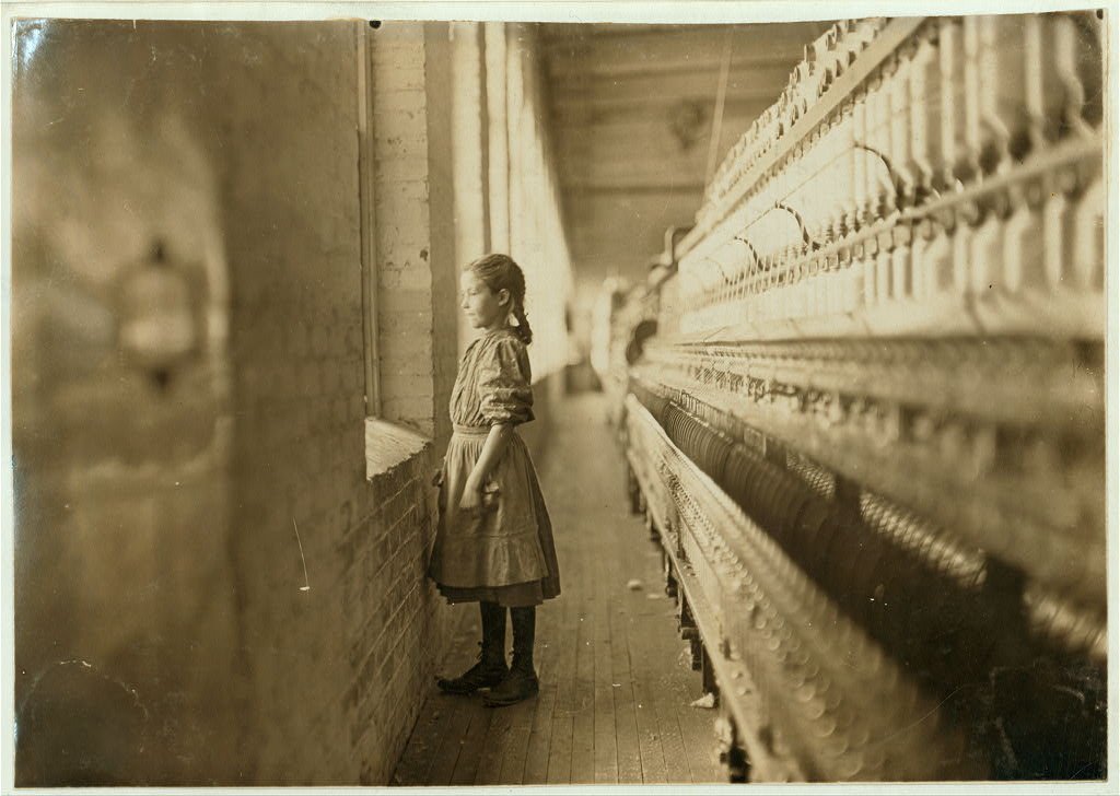 A young girl in a vintage dress stands by a brick wall, looking out a window in a textile mill with large weaving machines along the right side.