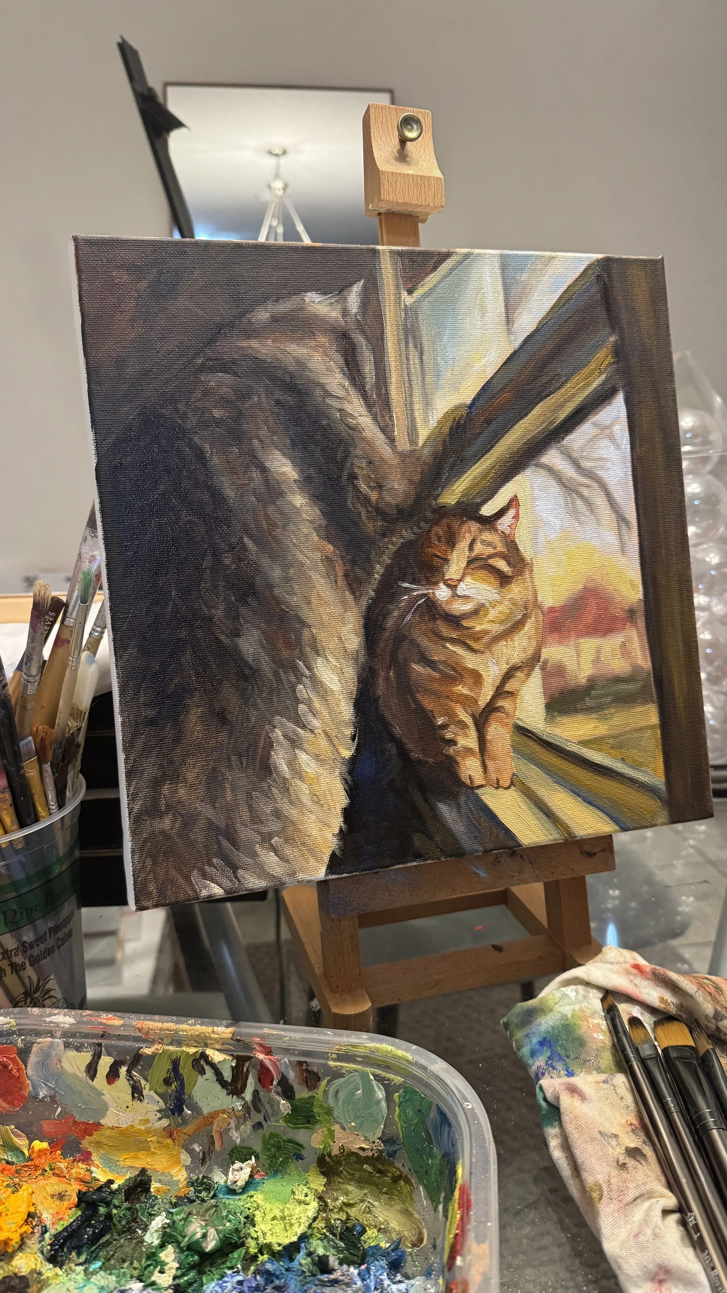 Painting of a cat looking out of a window, with a tree and a sunset in the background. The painting is on an easel with painting supplies nearby.
