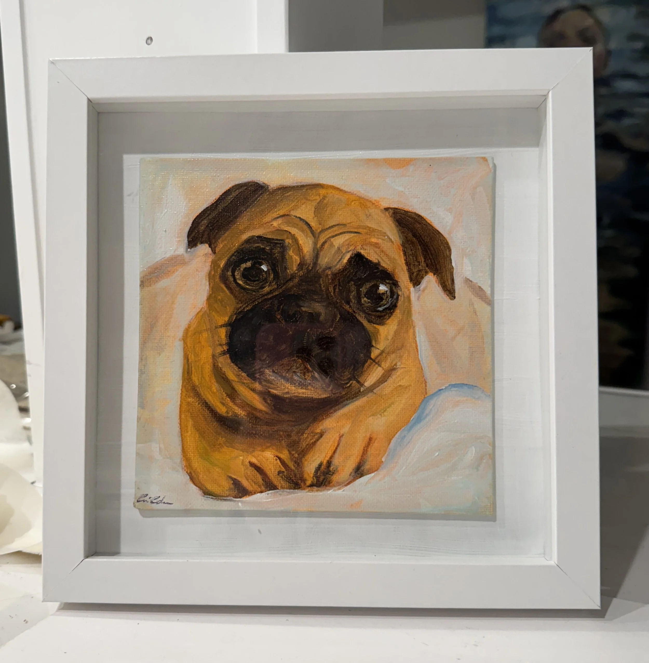A framed oil painting of a pug dog with large dark eyes, painted in warm tones with a cream background, signed in the bottom left corner.
