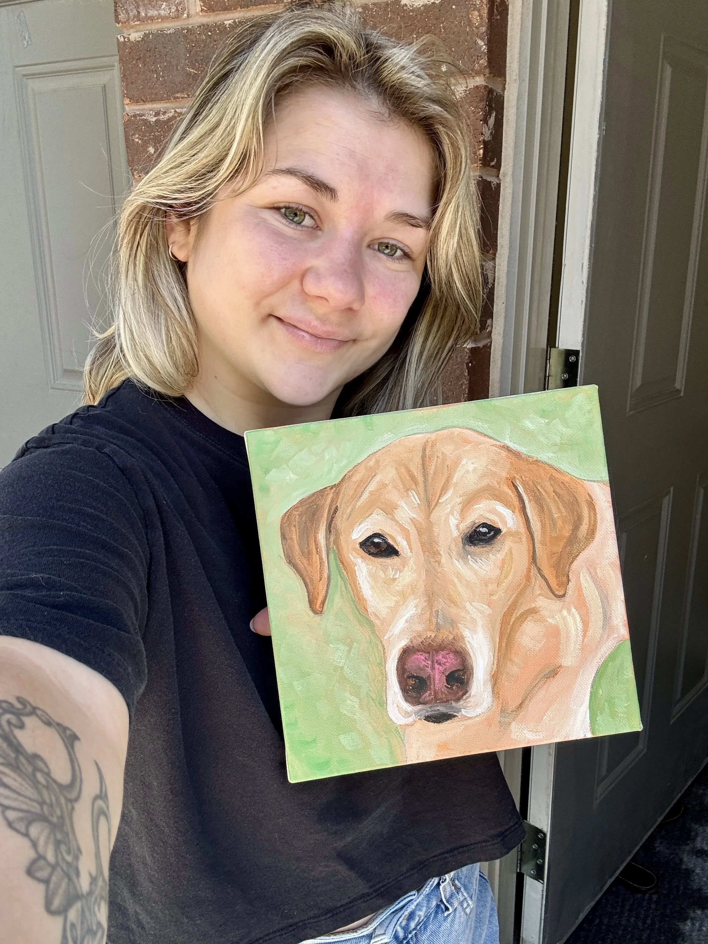 A young woman with blonde, wavy hair takes a selfie while holding a colorful painting of a dog. She is standing outside near a brick wall and a door.