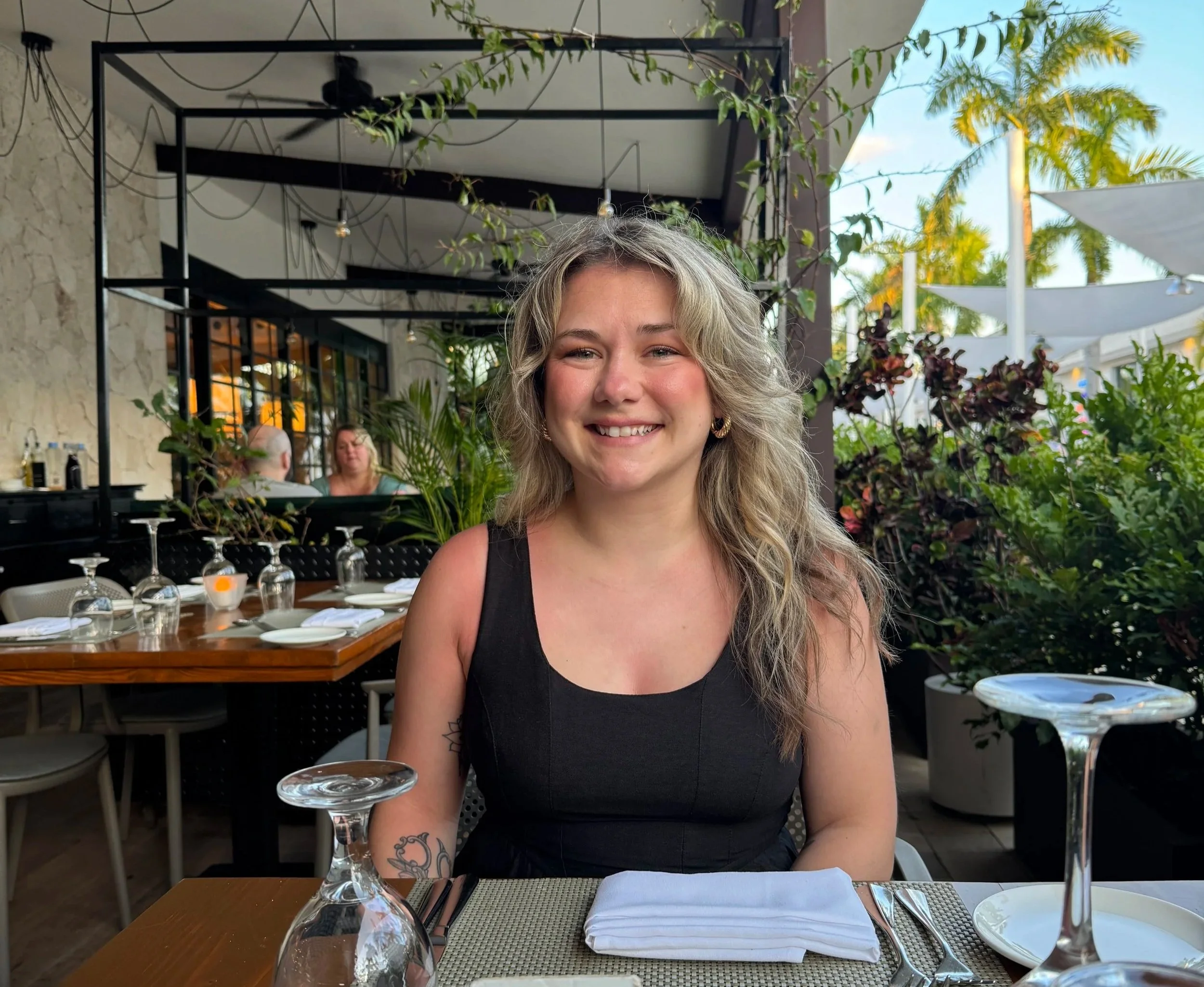 A young woman with blonde wavy hair and a black dress sits at an outdoor restaurant table, smiling. The table is set with wine glasses, napkins, and plates. There are plants and trees in the background, and a building with a stone wall and a large window.