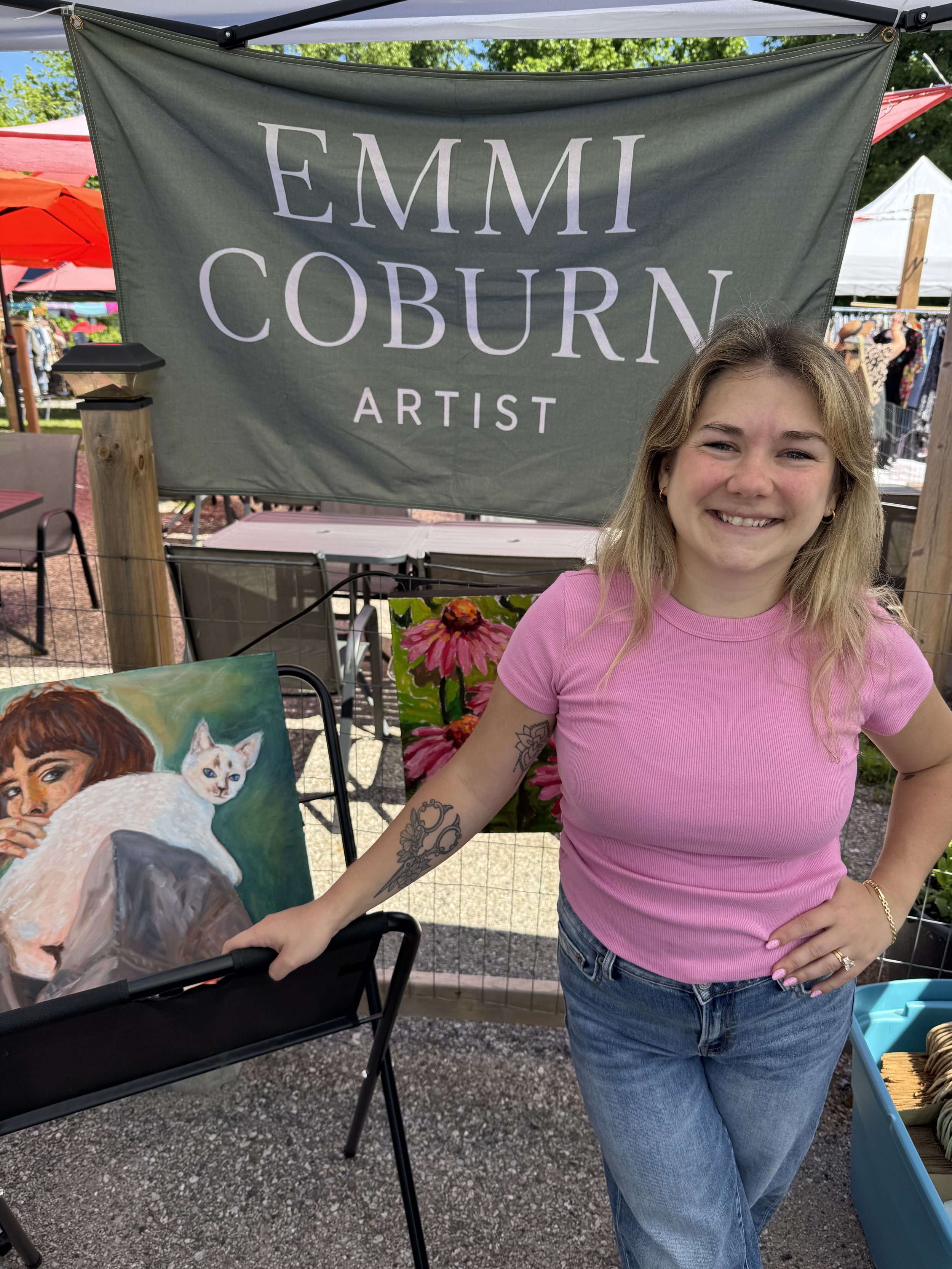 Emmi standing at an outdoor art market, wearing a pink shirt and jeans, with a painting of a woman and a white cat in front of her and art booths and tents in the background. A sign in the background reads 'EMMI COBURN ARTIST'.