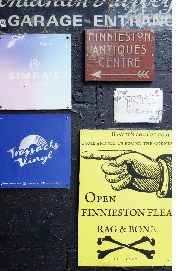 Several signs on a black brick wall, including a red sign for Finneston Antiques Centre pointing left, a white sign with non-Latin script, and a yellow flyer advertising a flea market at Finneston, with a graphic of a pointing hand and crossed bones.