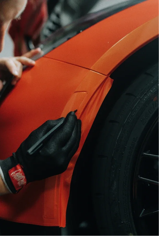 A person wearing black gloves is using a tool to apply orange protective film to a car's body near the wheel.