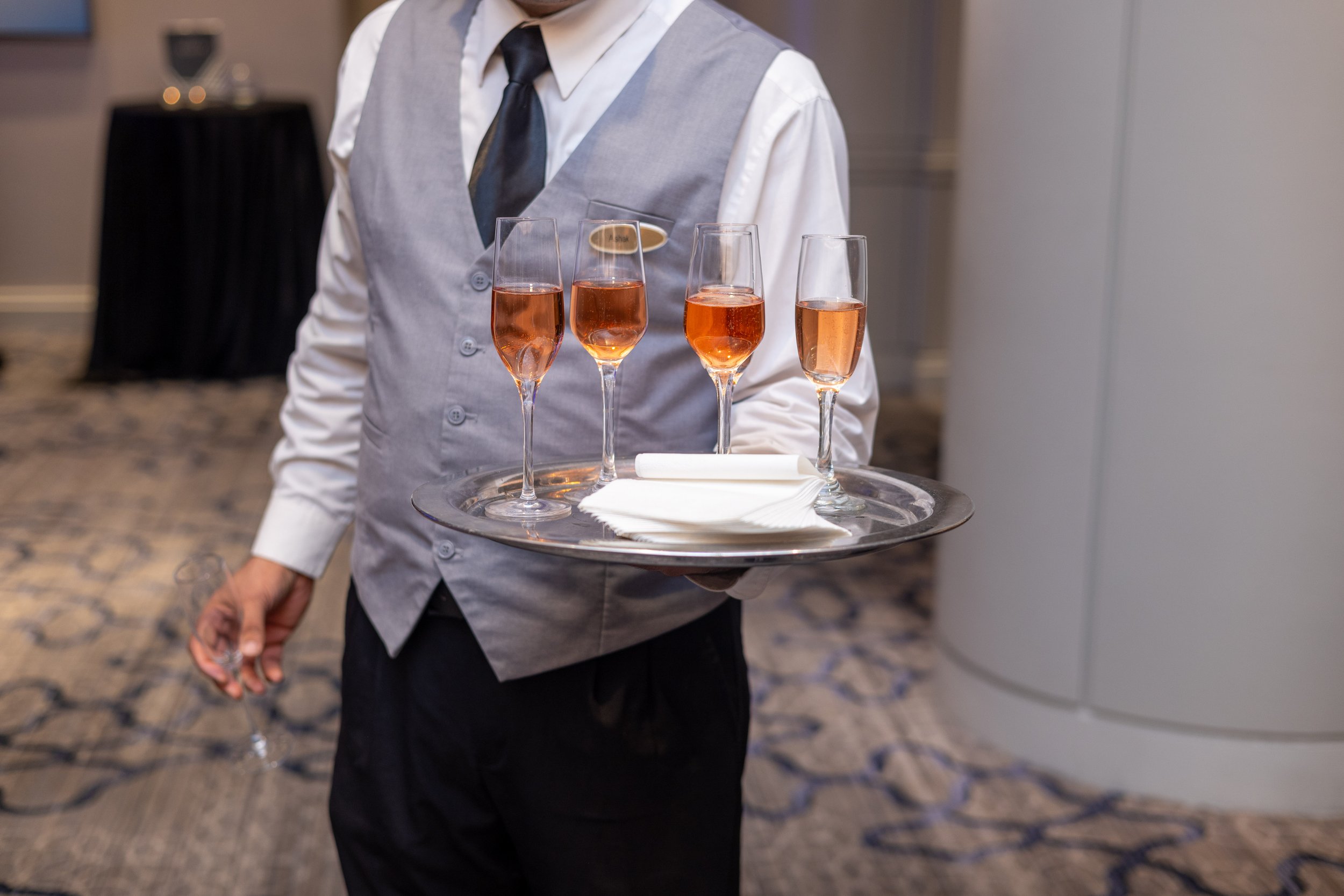 A waiter dressed in a white shirt, grey vest, black vest, and black tie holding a tray with four glasses of pink champagne.