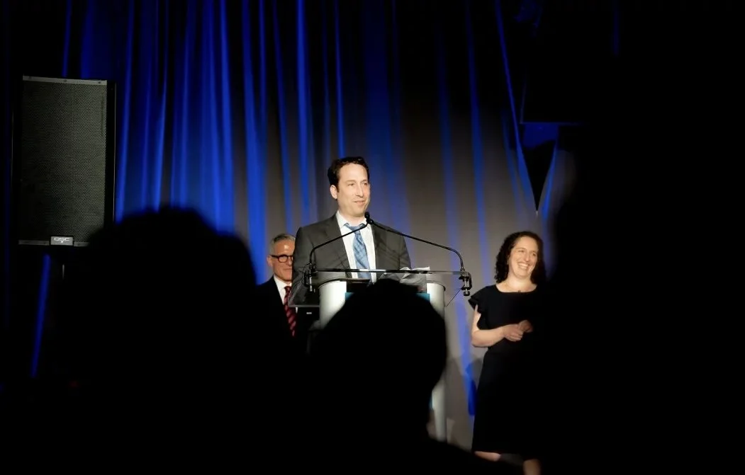 Man in suit speaking at a podium on stage with blue curtains, a woman in black smiling beside him, and a man with glasses watching behind.