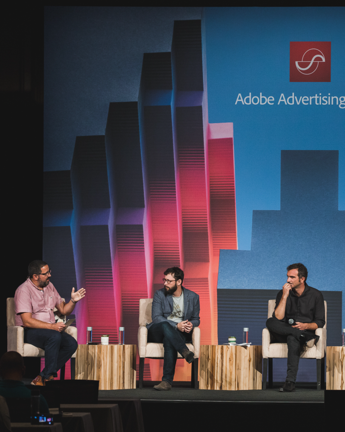 Three men sitting on stage in front of a large screen displaying the Adobe Advertising logo and colorful graphics, engaged in conversation during a panel discussion.