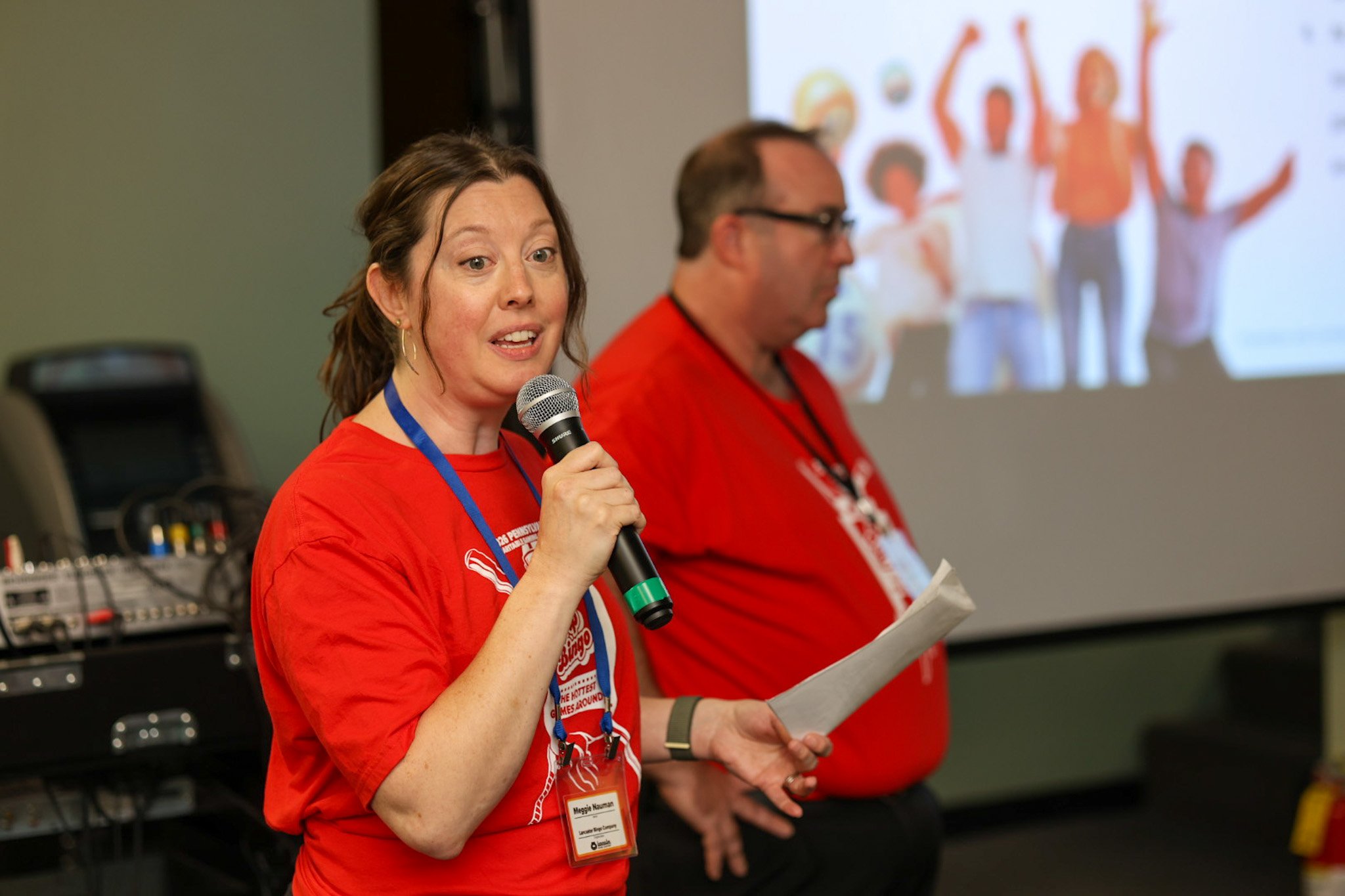 Woman speaking into a microphone at an indoor event, with a man standing beside her and a large screen displaying a blurred image of people with raised hands in the background.