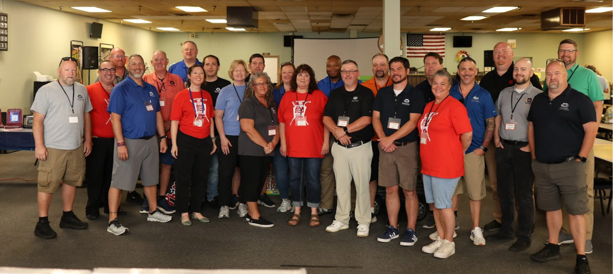 Group of people posing for a photo indoors, some wearing red, blue, and black shirts with badges, with American flag in the background.