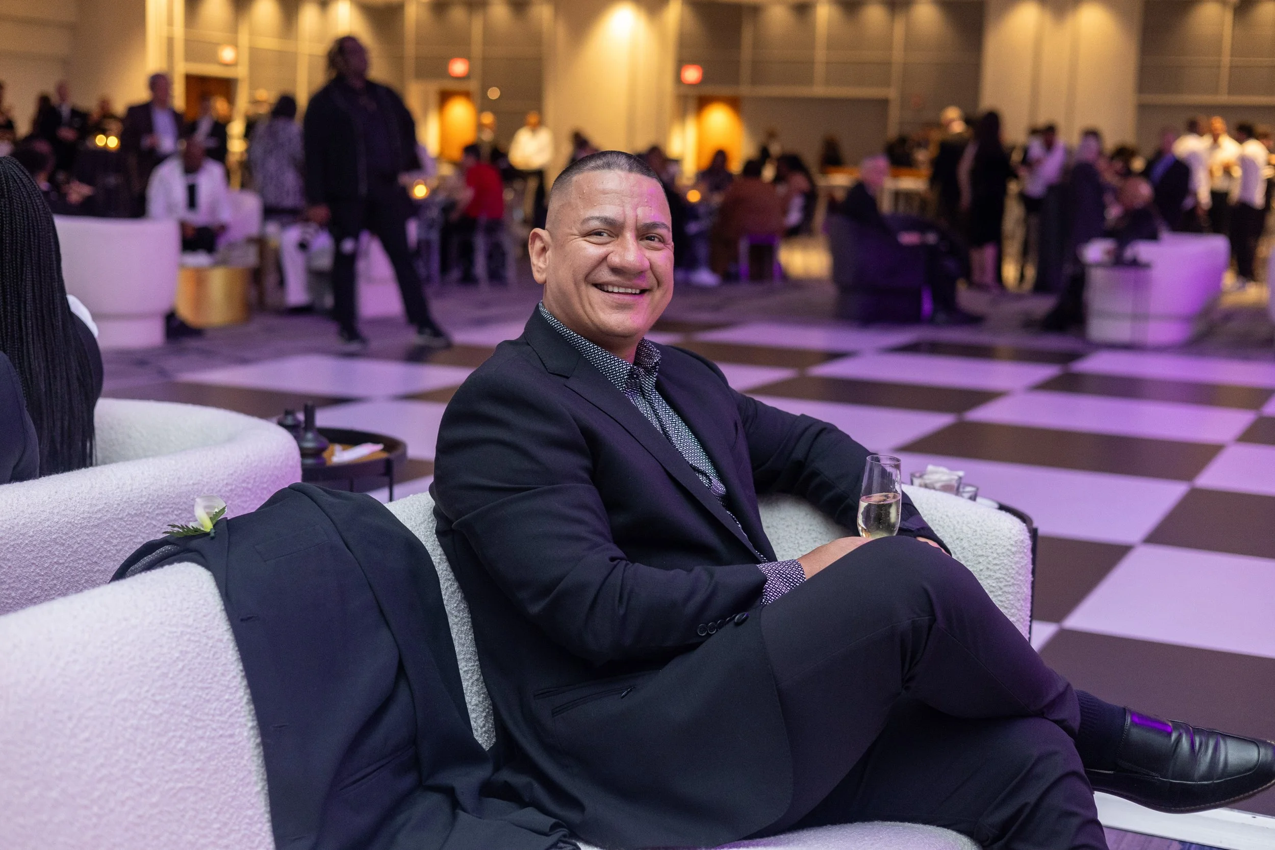 A man in a dark suit smiling while sitting on a white sofa at a social event, with a glass of champagne in his hand, in a large decorated hall filled with other guests.
