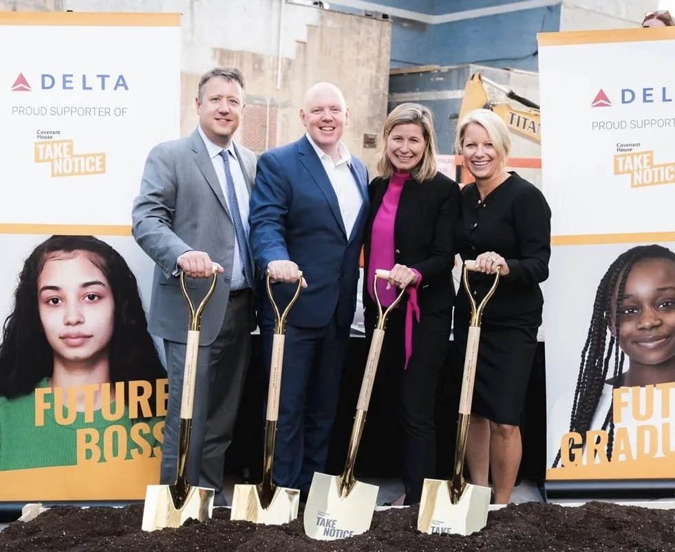 Four people in business attire holding golden shovels, standing in front of banners supporting Delta Airlines and a community project called 'Covenant House Take Notice.' The event appears to be a groundbreaking ceremony with the ground covered in da