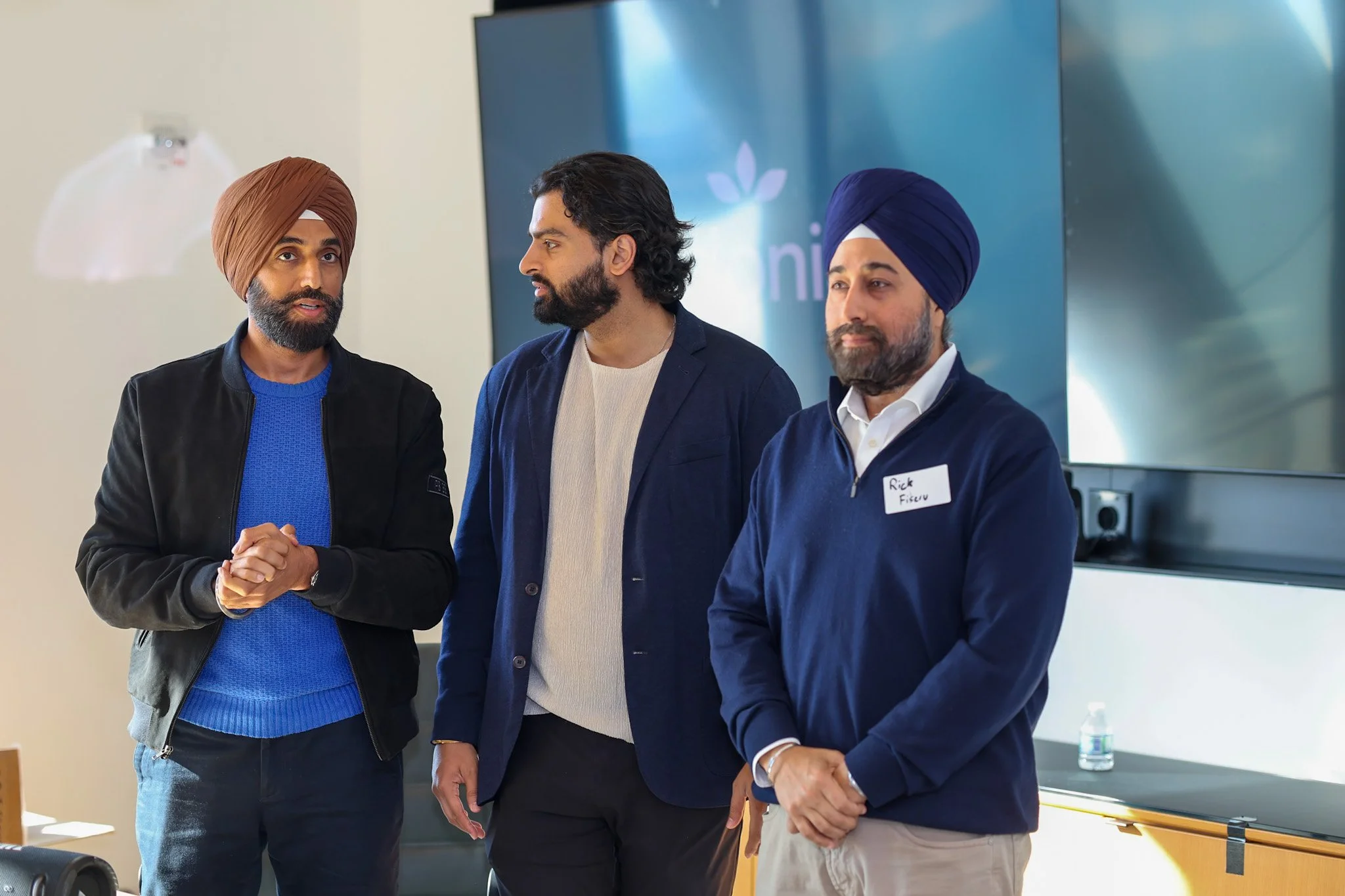 Three men standing and speaking in a conference room; two are wearing traditional Sikh turbans and the third is with curly hair and a beard; they appear to be engaged in a discussion.