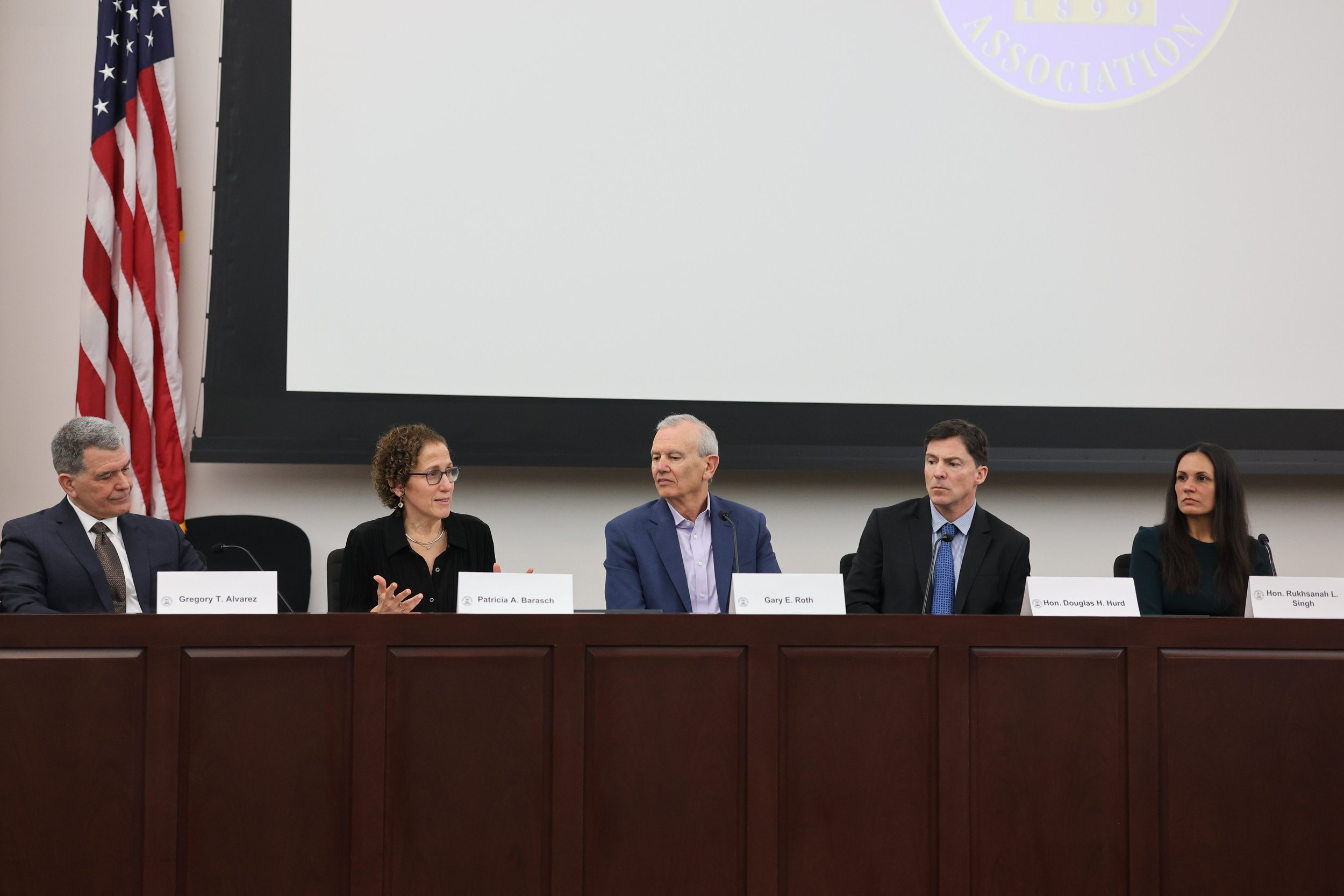 A group of five people sitting at a conference table with name tags in front of them, participating in a panel discussion in a formal setting. There is an American flag on a pole to the left and a blank projection screen behind them.