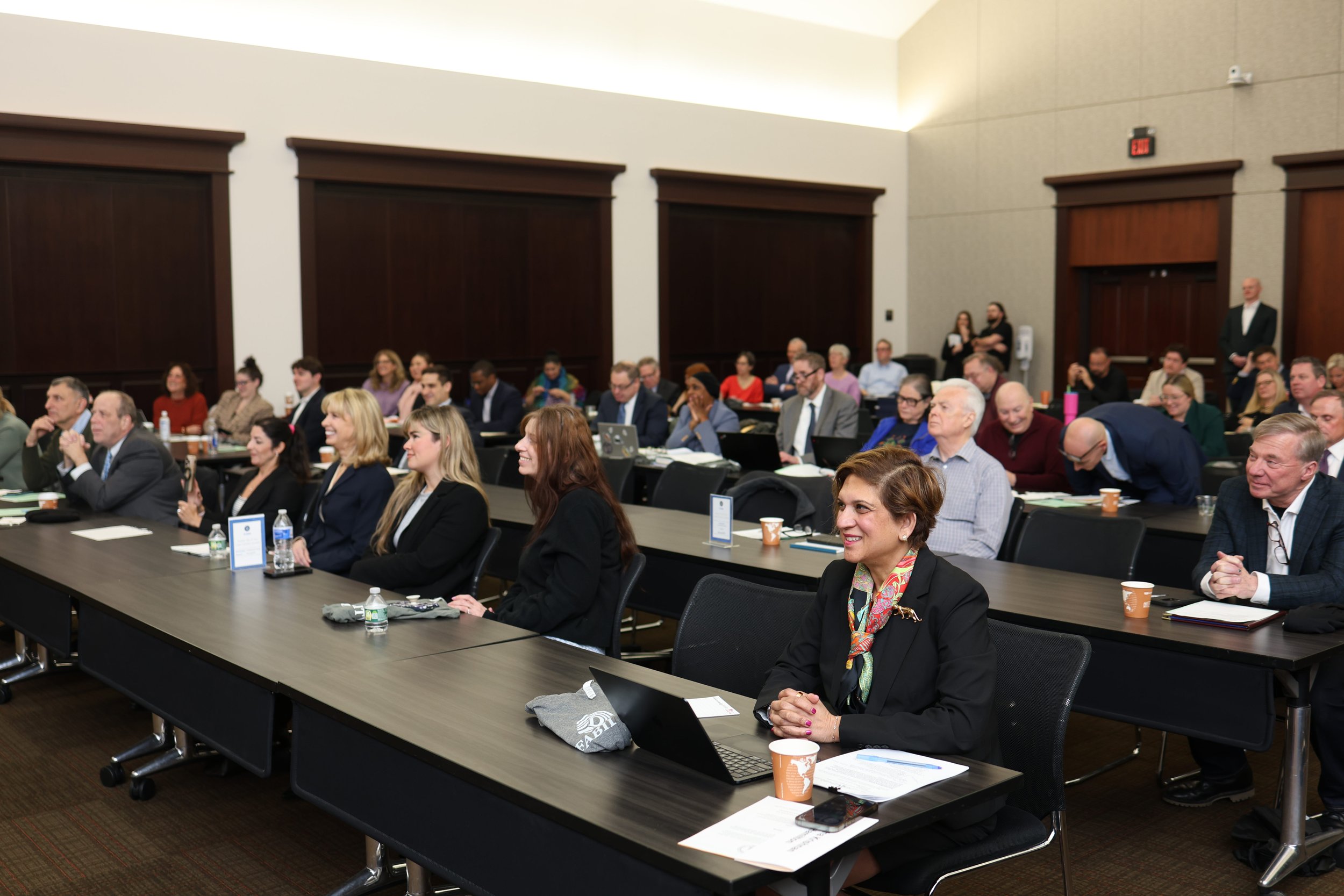 People attending a professional conference in a large room, seated at long tables with laptops, notebooks, and coffee cups.