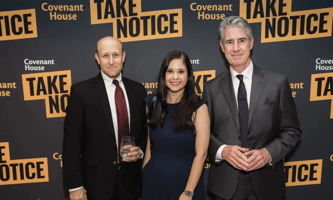 Three people in formal attire standing in front of a backdrop with the text 'Covenant House' and 'Take Notice'. The man on the left is holding a glass of water, the woman in the middle is smiling, and the man on the right has his hands clasped.