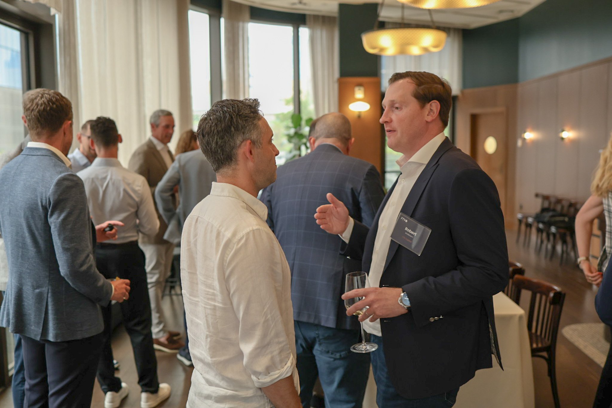 Two men having a conversation at a social gathering or networking event, with several other people in the background in a well-lit, modern room.
