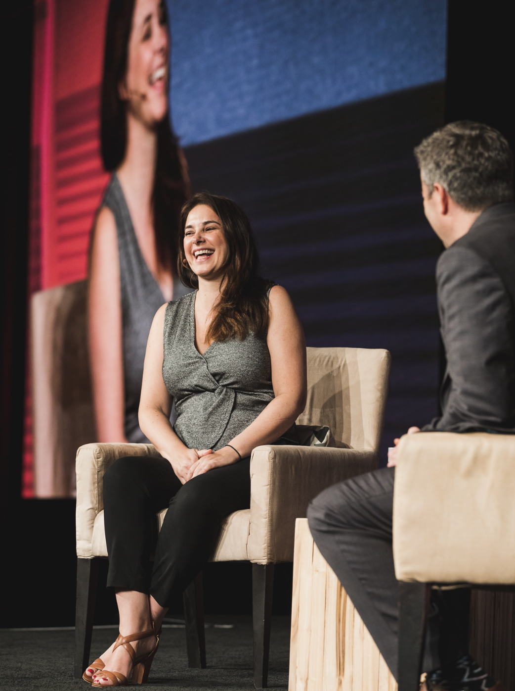 A woman with long brown hair, wearing a gray sleeveless top and black pants, sitting on a beige chair, smiling and laughing during a talk show or interview. A man in a gray suit, sitting opposite, is partially visible. A large screen behind them show