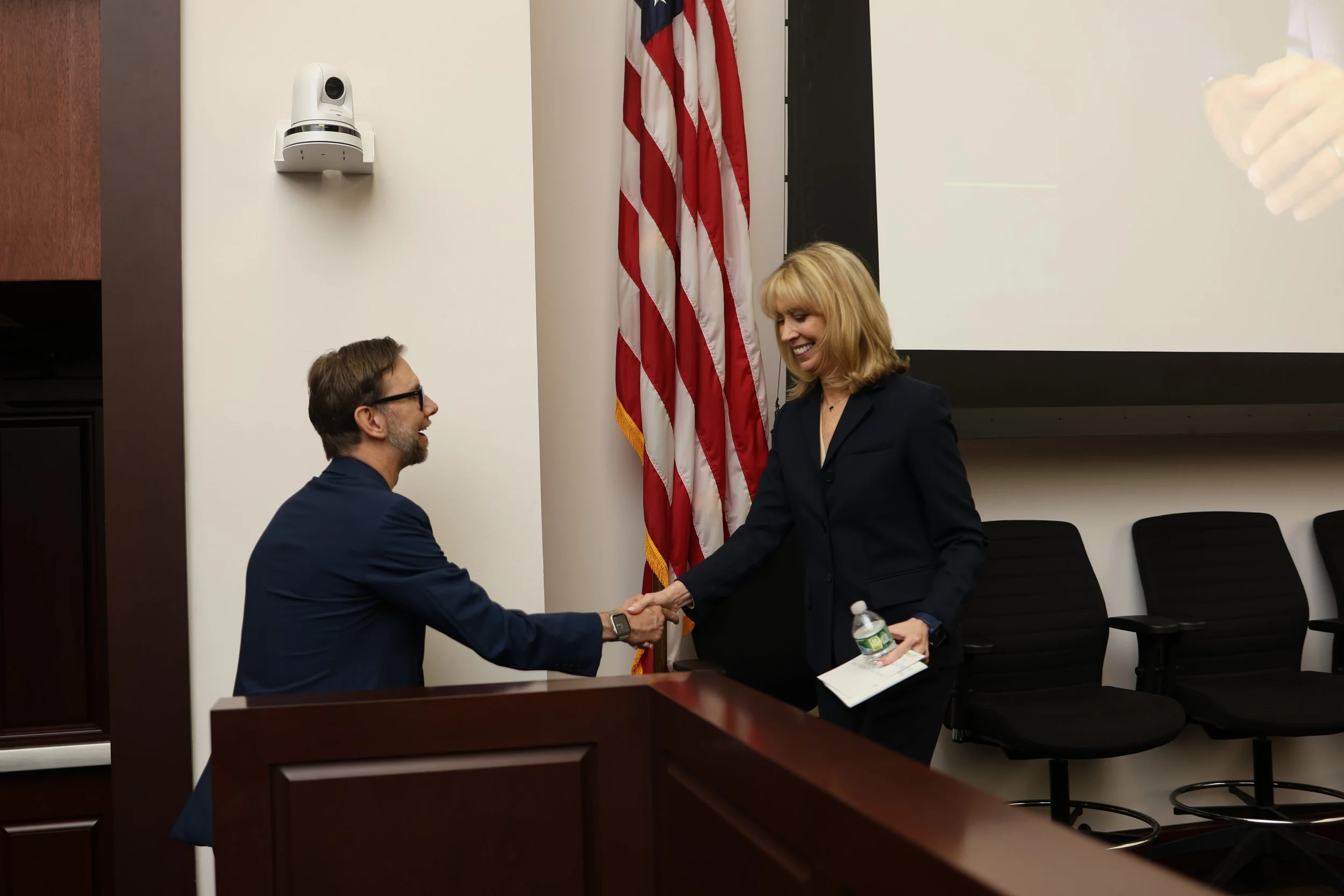 A man and woman in business attire shaking hands in a conference room. The woman is smiling and holding a water bottle and papers, while the man is smiling and wearing glasses. An American flag is visible behind them, and there is a large screen and 