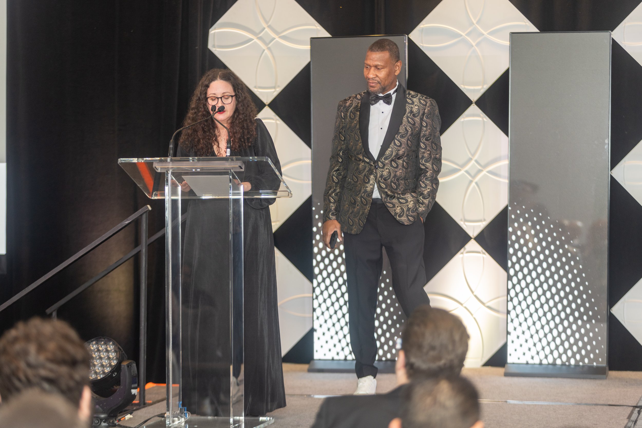 A woman with curly hair and glasses speaking at a clear podium, and a man wearing a black tuxedo with a patterned jacket standing beside her on a stage with geometric black and white background panels.