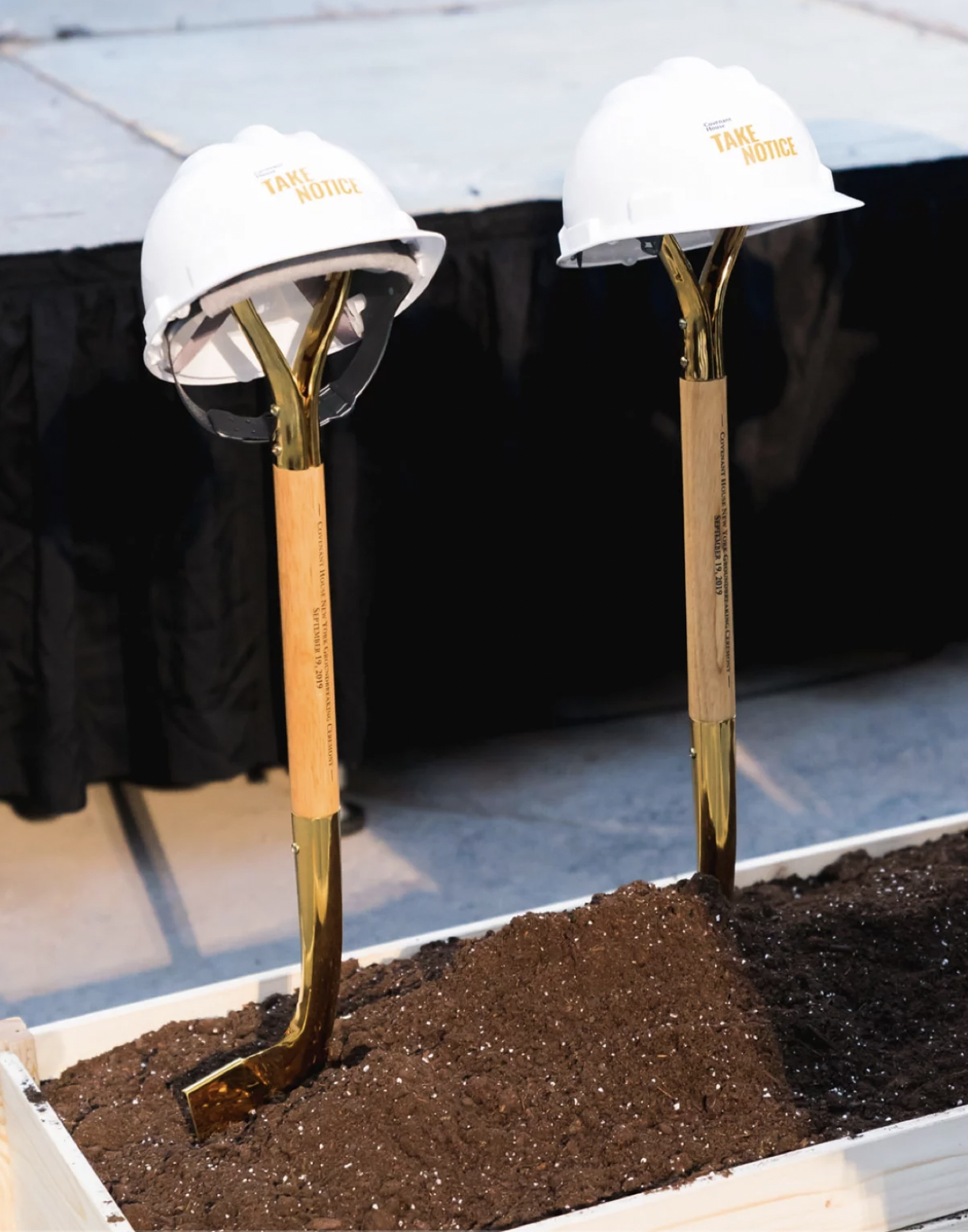 Two construction shovels with white safety helmets resting on the handles, placed in a flower bed with dark soil, at a construction site.