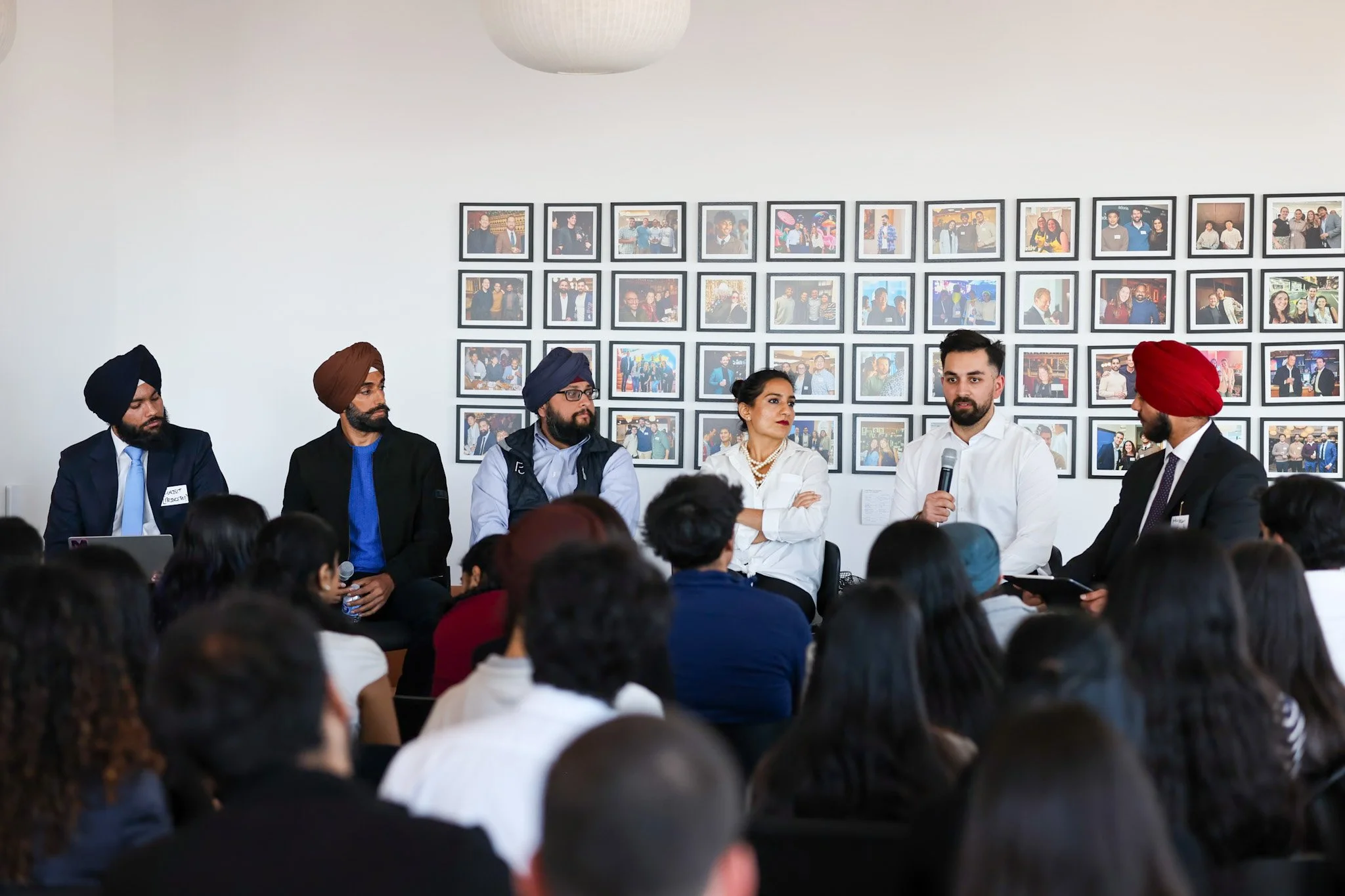 Panel of five diverse individuals seated at a table speaking to an audience with framed photographs on the wall behind them.