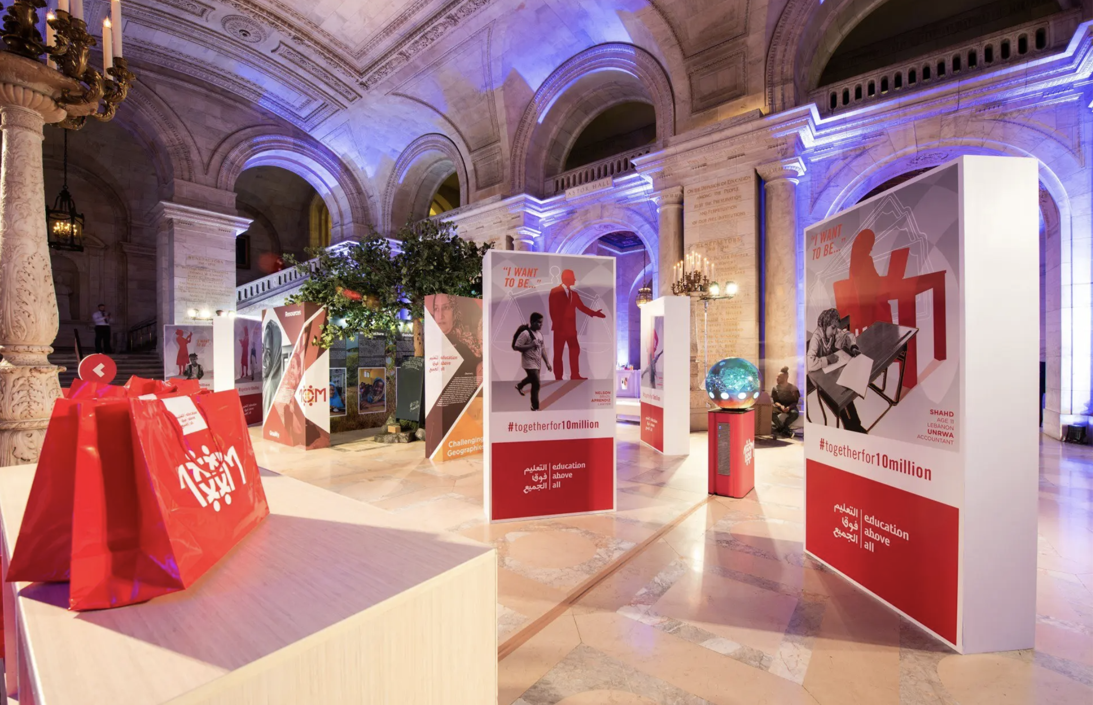 Exhibition in a historic hall with high arches and ornate columns, featuring informational posters about education and career aspirations, a globe, and red shopping bags with a logo on a table in the foreground.
