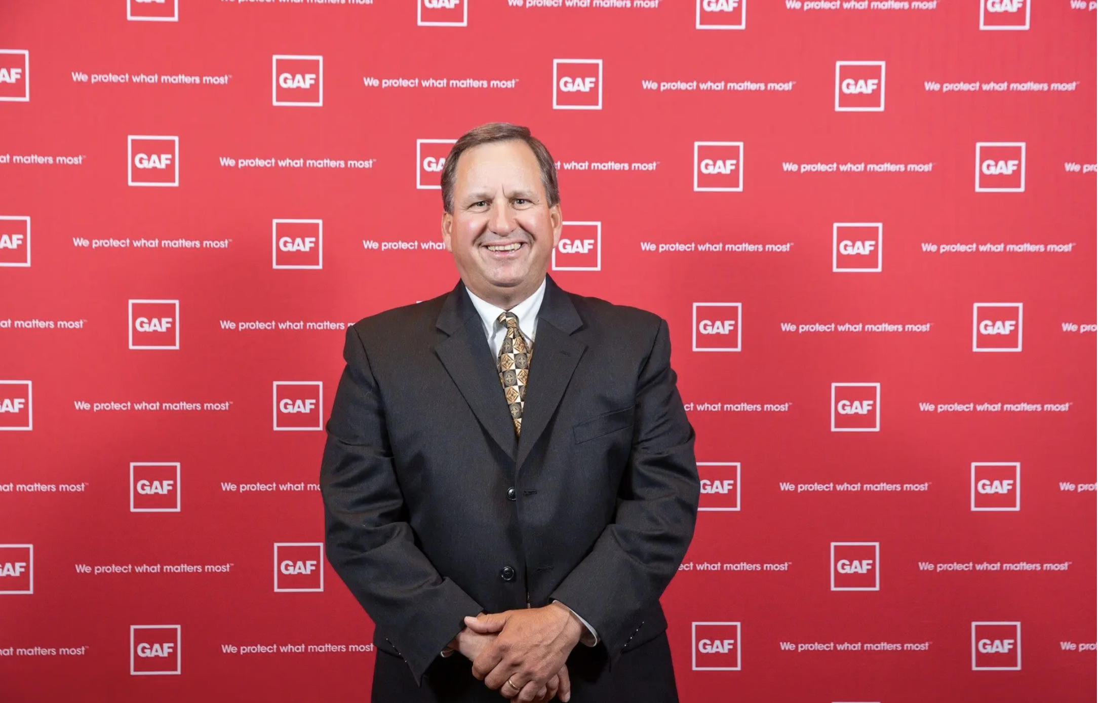 A smiling man in a dark suit and patterned tie stands in front of a red backdrop with white logos and text that read "GAF" and "We protect what matters most."