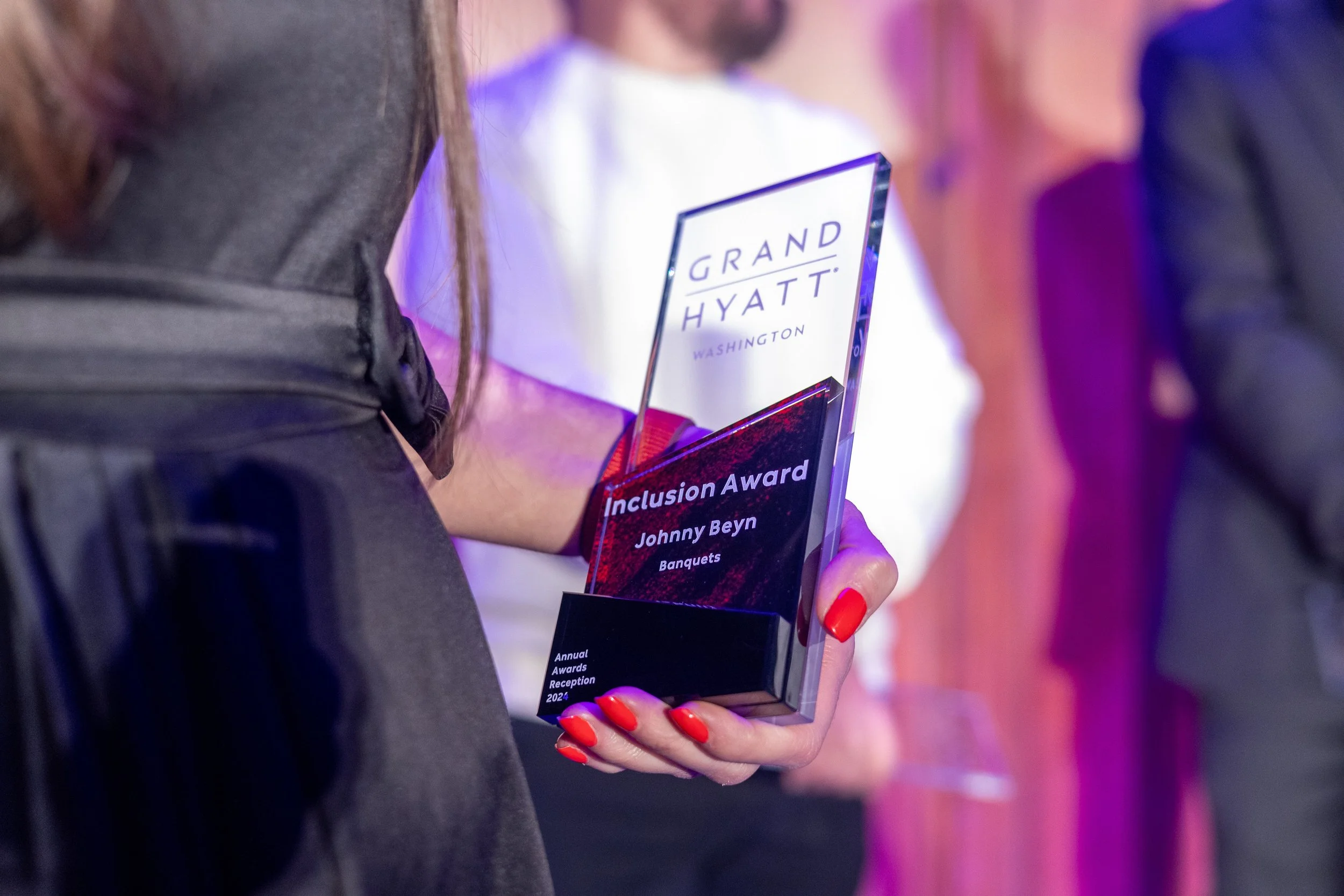 Person dressed in black holding a clear award with the Grand Hyatt Washington logo, receiving an Inclusion Award for Johnny Beyn at an event.