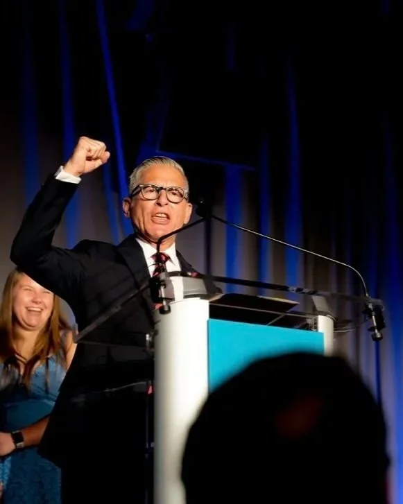A man in a suit and glasses is speaking at a podium, raising his fist in a gesture of triumph or solidarity, with a woman smiling behind him.