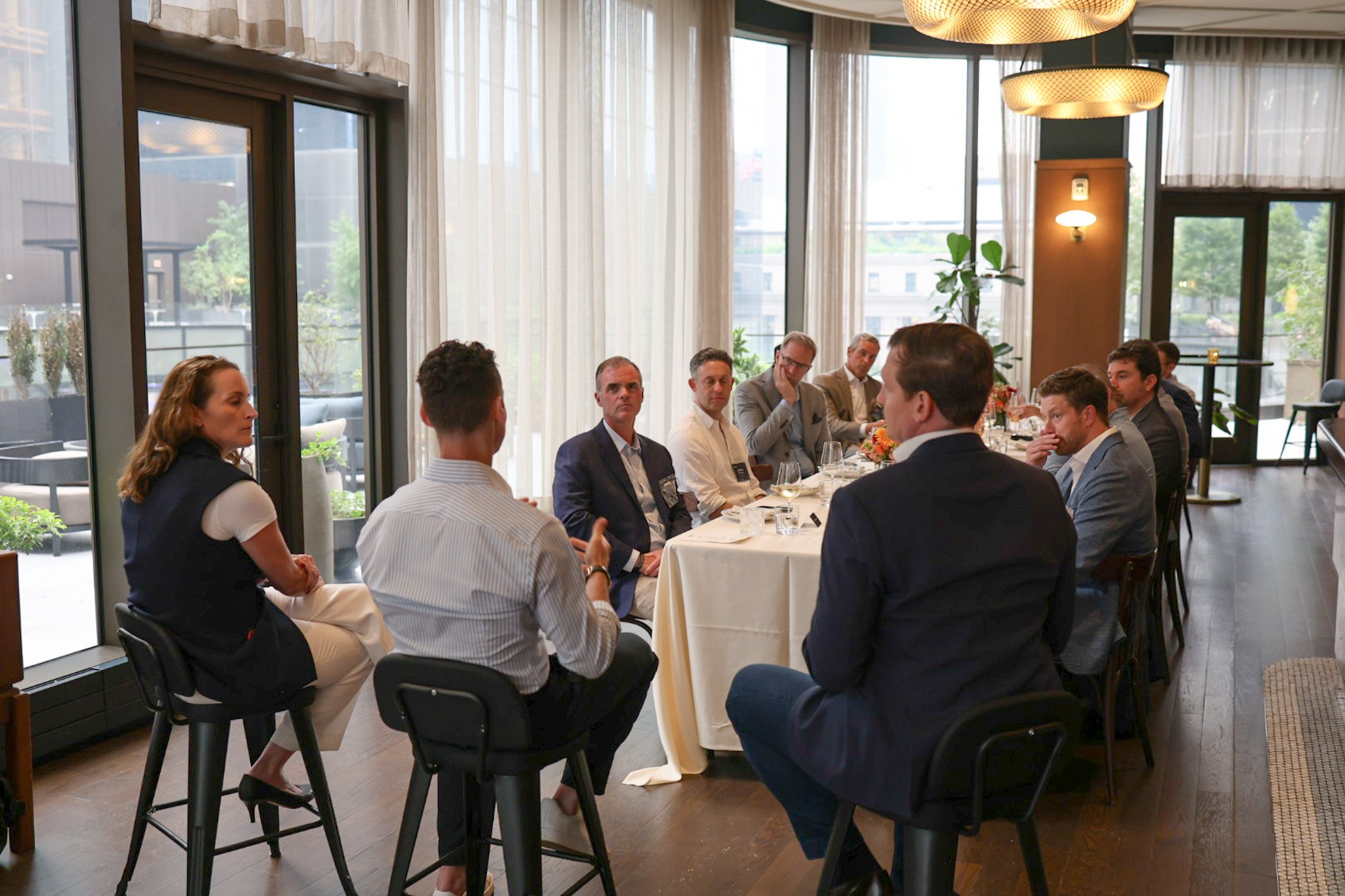 A group of professionals in business attire sitting around a table in a brightly lit restaurant or conference room with large windows and sheer curtains, engaged in a discussion.
