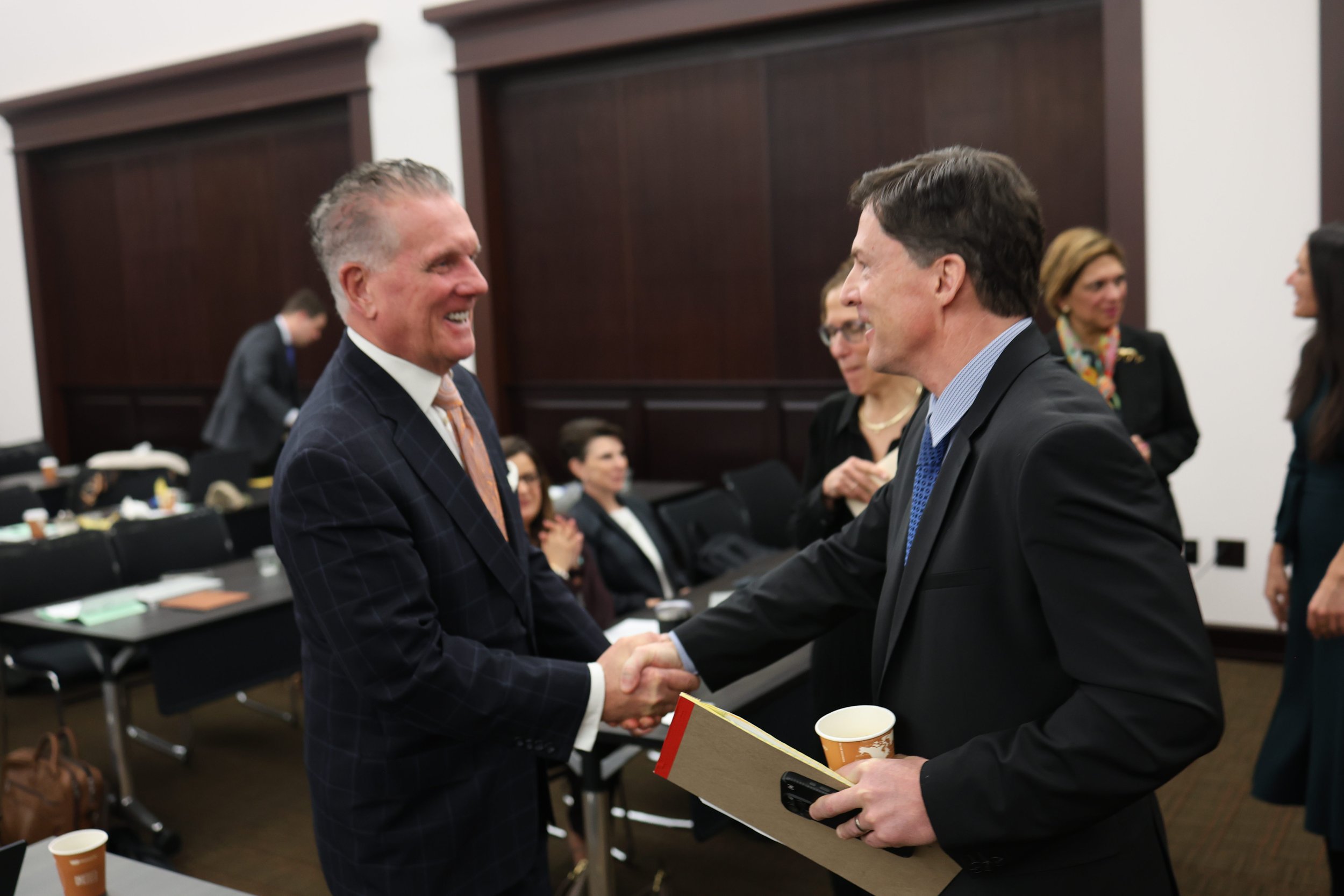 Two men in suits shaking hands in a formal setting, surrounded by women and men attending a meeting or event.