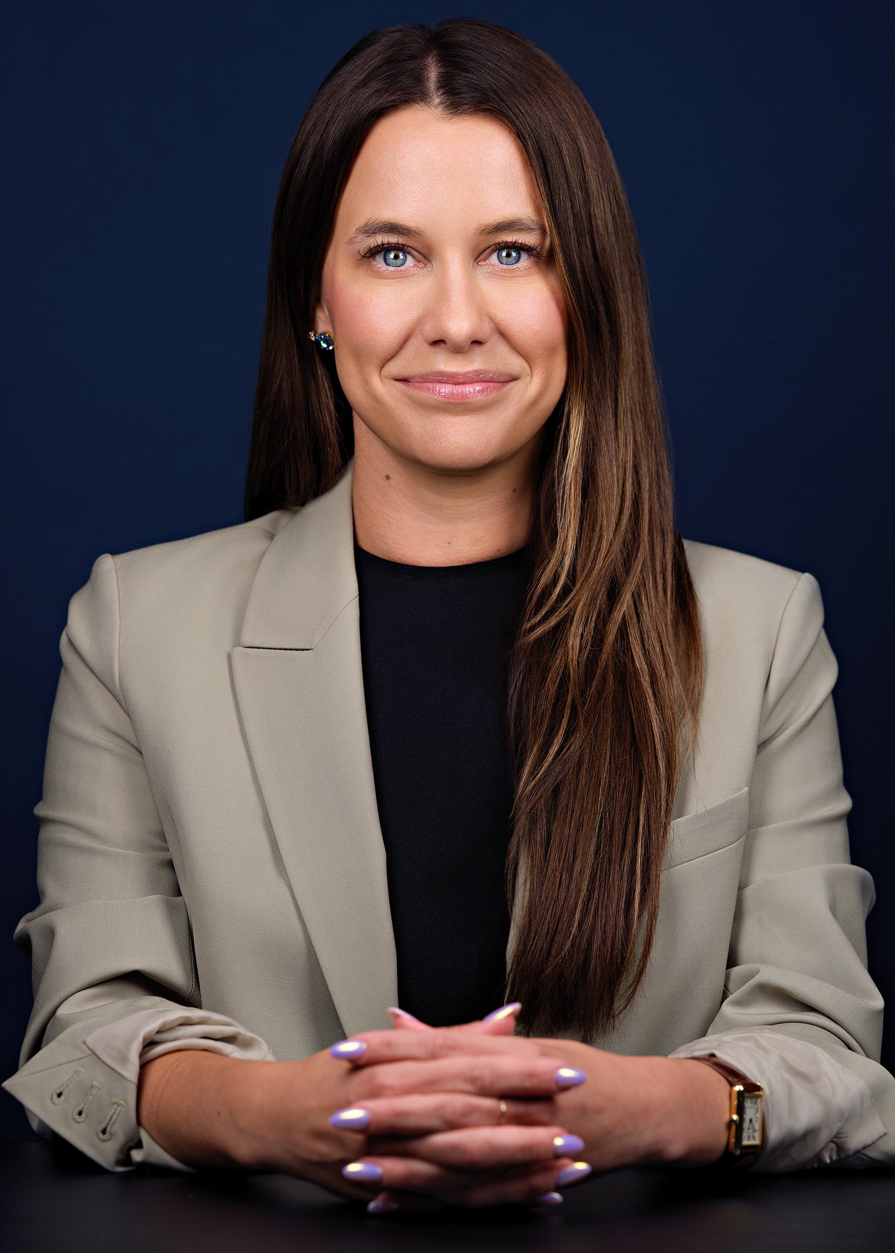 A woman with long brown hair and blue eyes sitting at a table with her hands clasped. She is wearing a beige blazer, black top, and has a gold watch on her wrist. She is smiling slightly against a dark background.