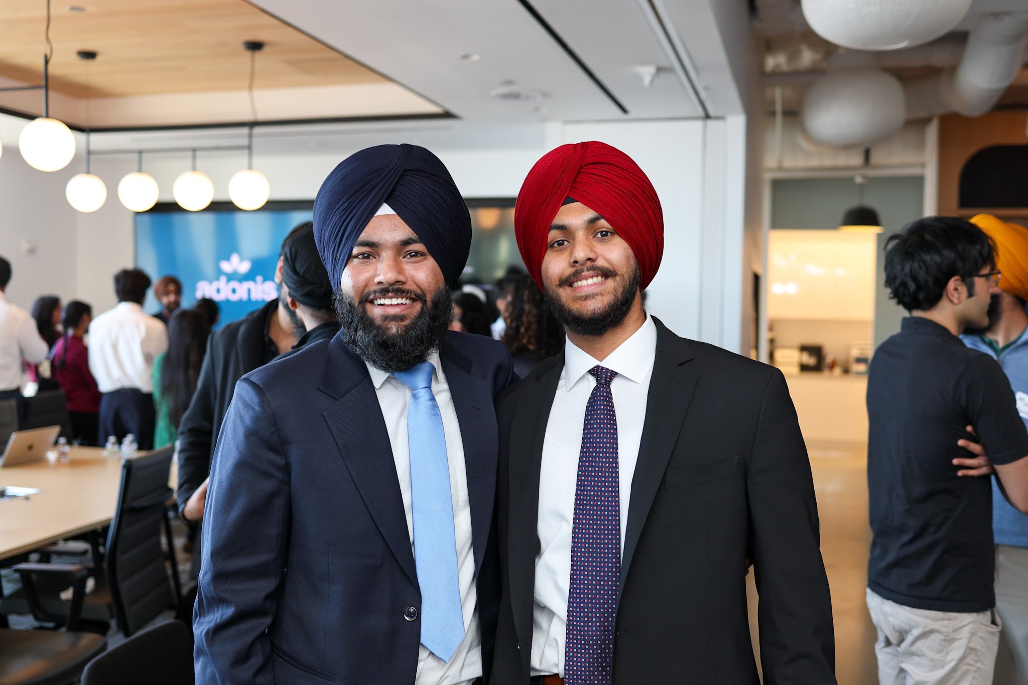 Two men wearing suits and turbans, one in navy and the other in red, smiling at a professional event or conference in a bright, modern room with other attendees in the background.