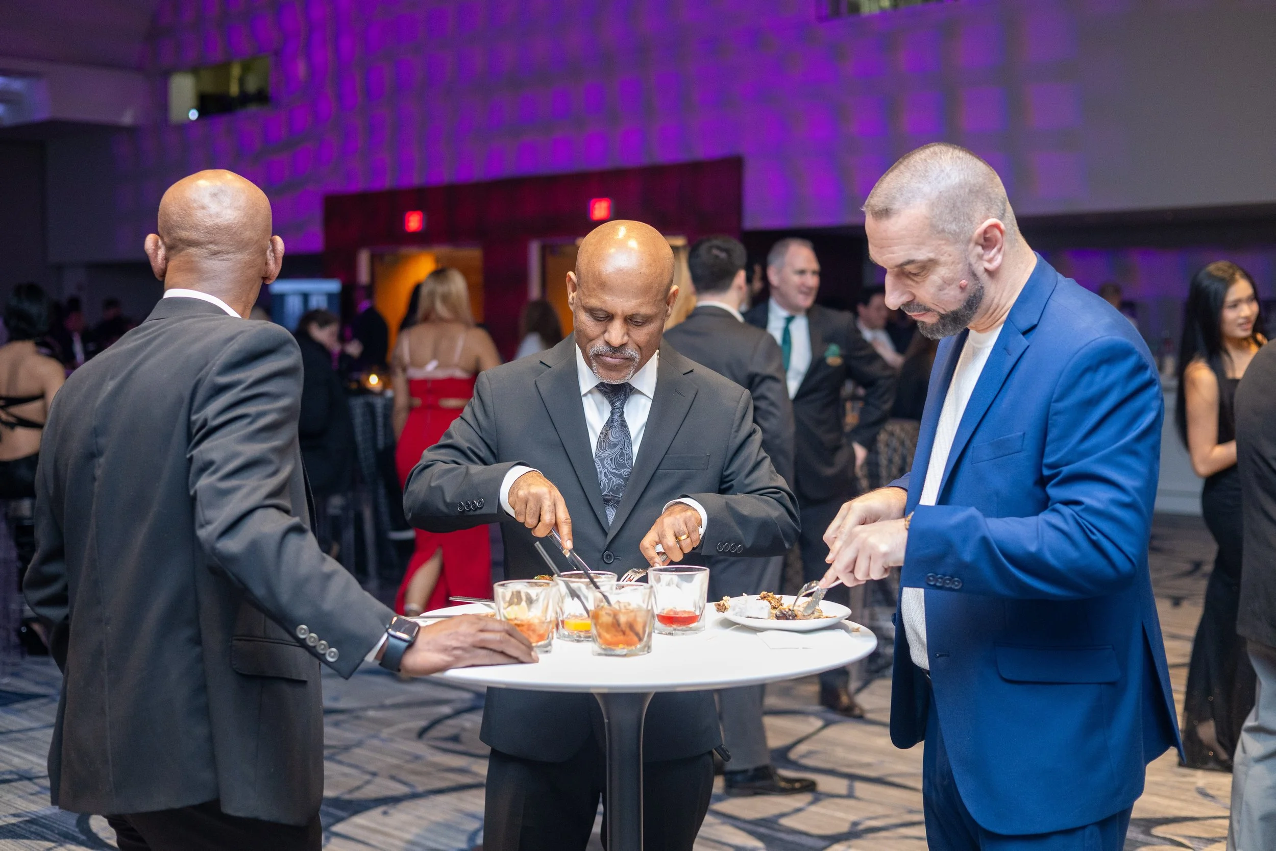Three men in formal attire at a buffet table at a social event, with one man serving himself food and the others looking on, in a dimly lit room with purple lighting and other guests in the background.