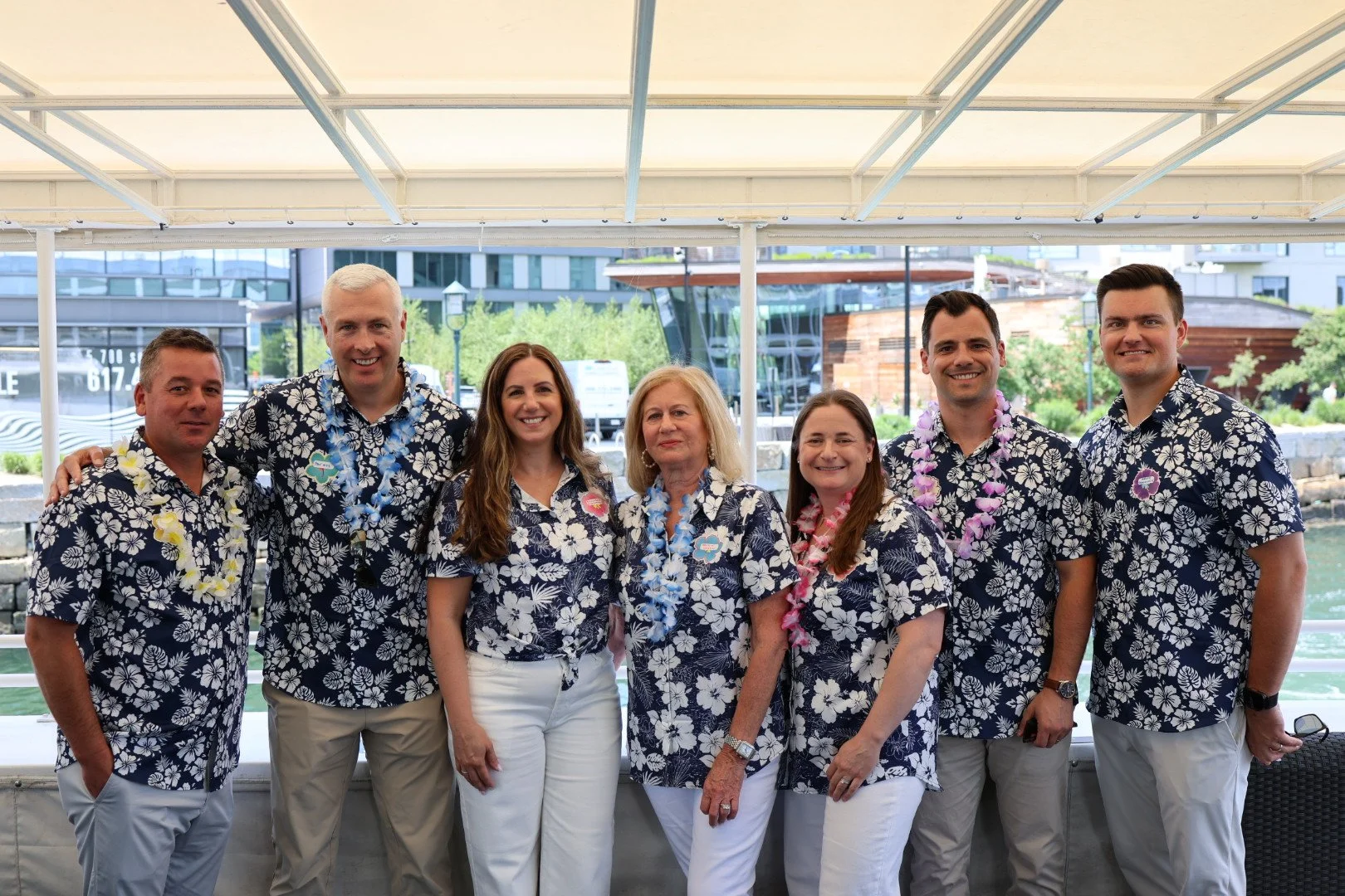 Group of seven people dressed in matching navy floral shirts and white pants, celebrating outdoors with leis, smiling, under a canopy with modern buildings and greenery in the background.