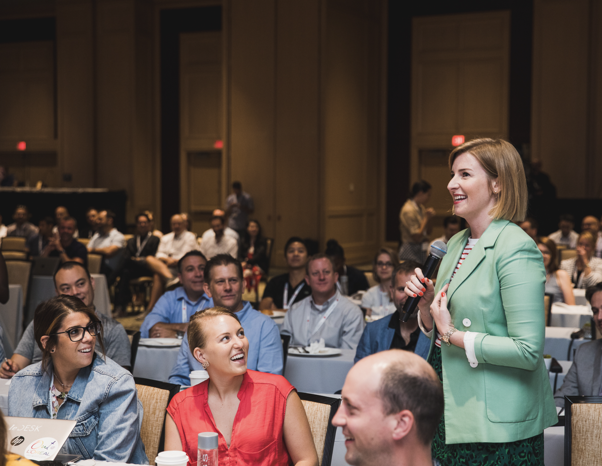 A woman in a light green blazer is speaking into a microphone during a conference, smiling and engaging with an audience that appears interested and attentive. The setting is a large, formal conference room with many audience members seated at tables