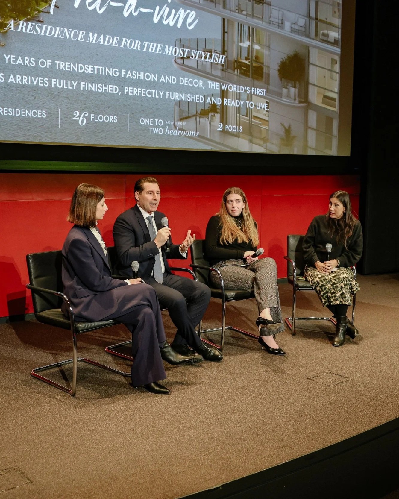 Four individuals seated in chairs on a stage during a panel discussion. The man second from the left is speaking into a microphone, wearing a dark suit and tie. The three women are also holding microphones. A large screen behind them displays an advertisement for a residence with details about floors, bedrooms, and pools.