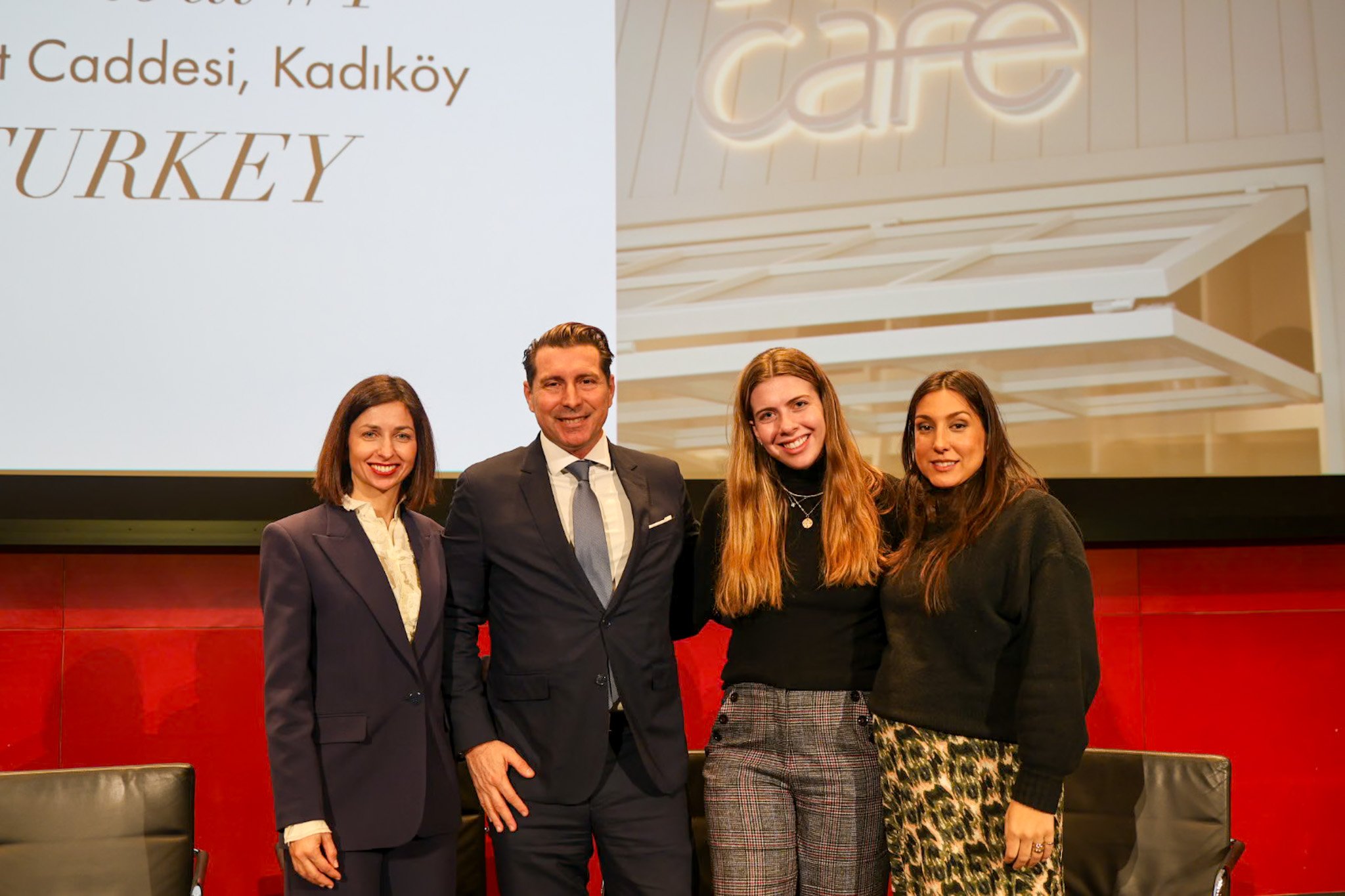 Four people standing together in front of a large screen displaying text about Istanbul, Kadıköy, Turkey, with a lit sign that says 'cafe' above them. They are dressed in business attire and smiling at the camera.