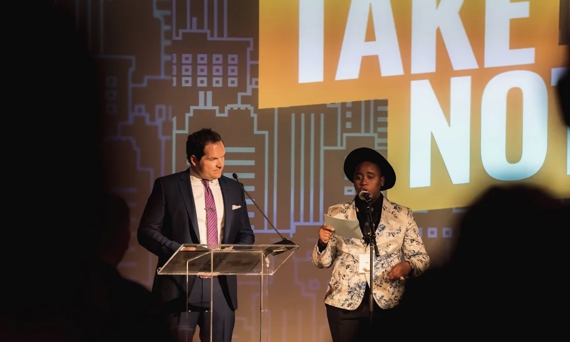 A man in a dark suit and purple tie stands behind a clear podium, looking at a woman speaking into a microphone. The woman is wearing a black hat and a floral blazer, holding notes. Behind them is a large screen with the words "TAKE NO" and a backgro