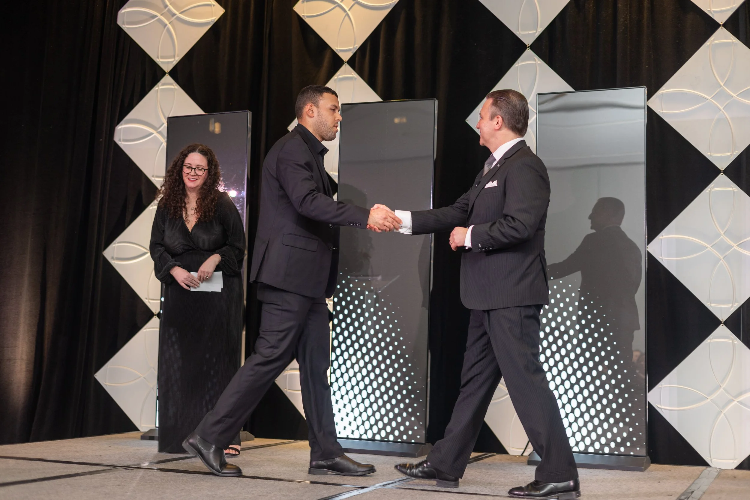 Two professionals shaking hands on a brightly lit stage during a corporate awards ceremony.