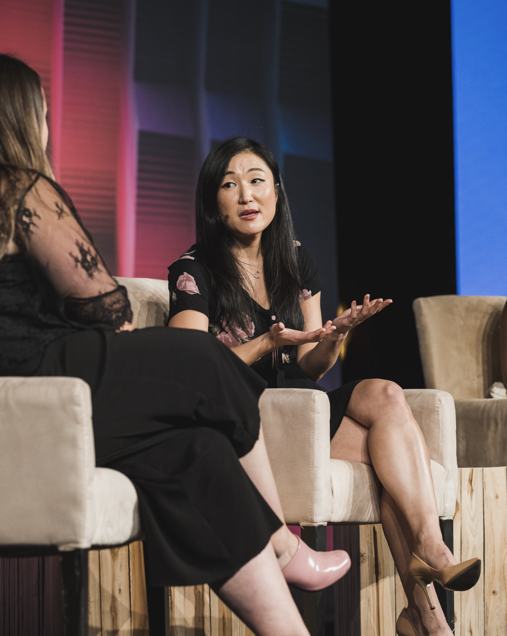 Two women having a conversation on stage, one speaking with hand gestures, seated on beige armchairs, with a colorful background.