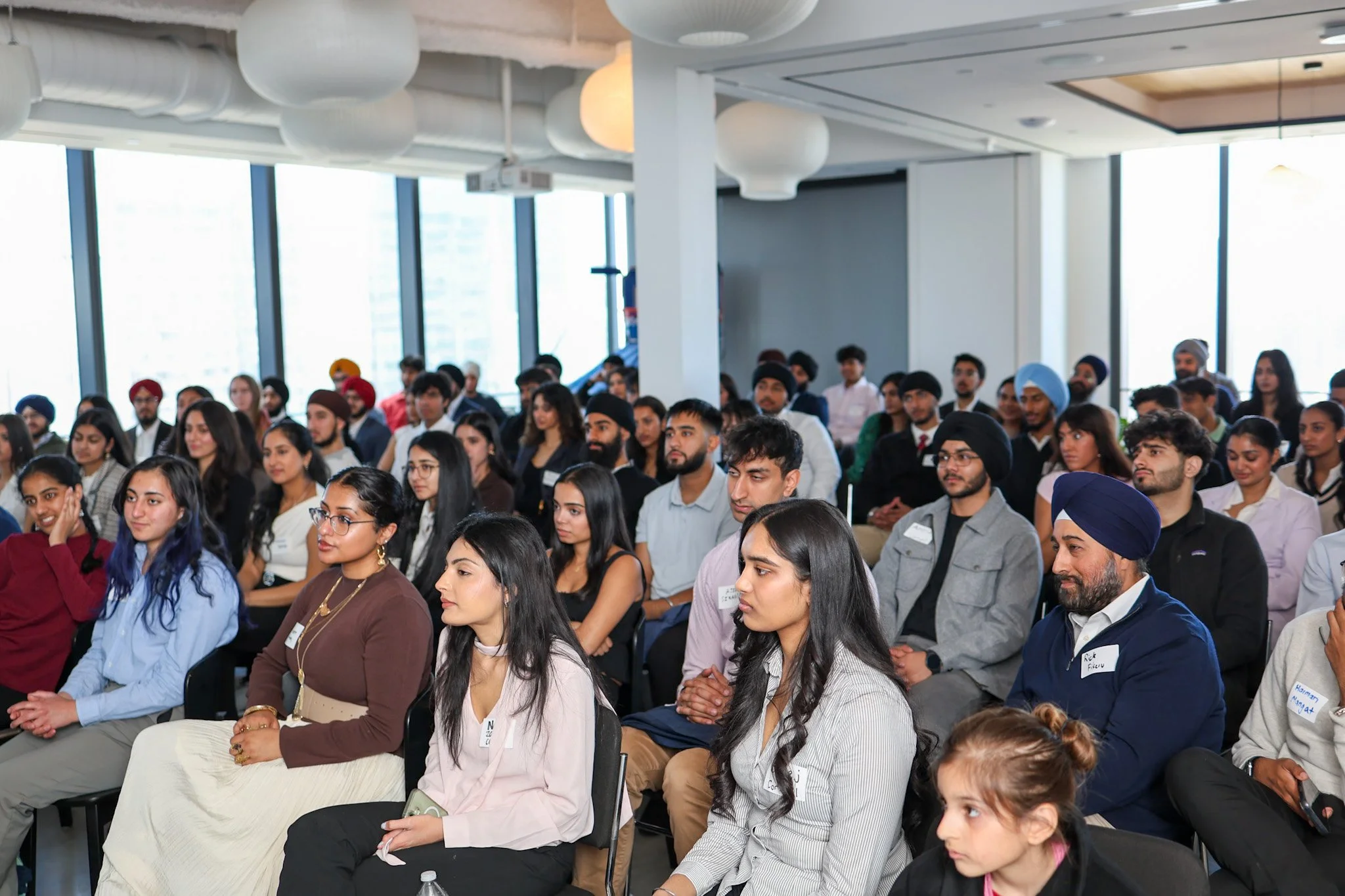 A large crowd of diverse people sitting attentively at a conference or seminar in a modern room with large windows and white decor.