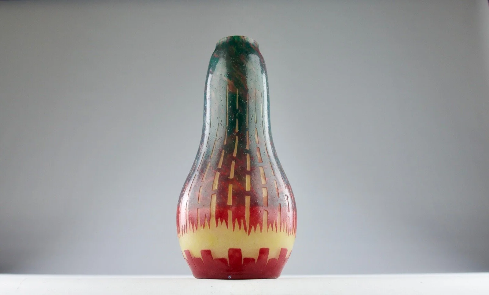 Colorful art glass vase with geometric patterns and gradient colors, placed on a white surface against a gray background.